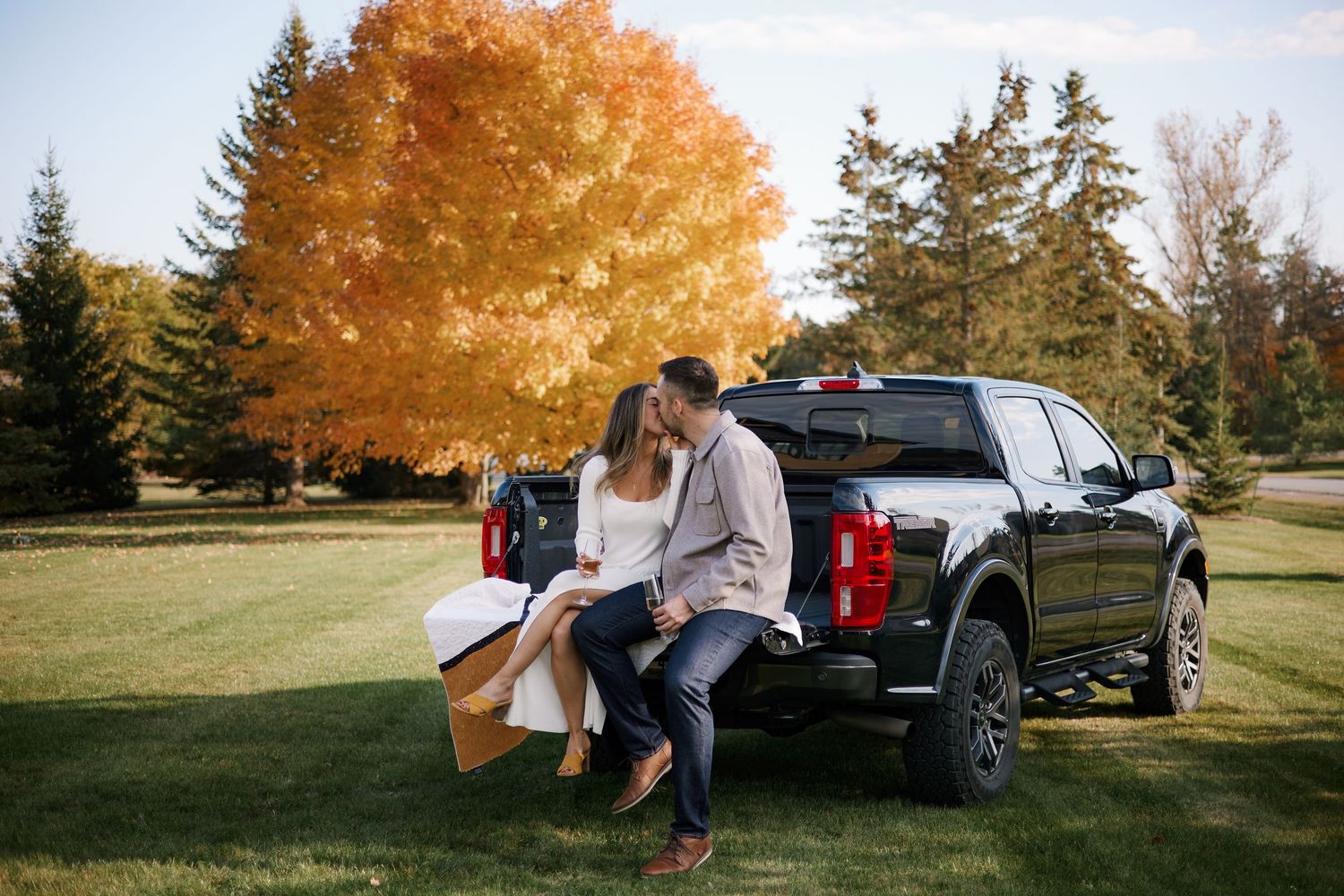 Couple sharing a romantic kiss on a black truck tailgate surrounded by vibrant autumn foliage in a scenic park.