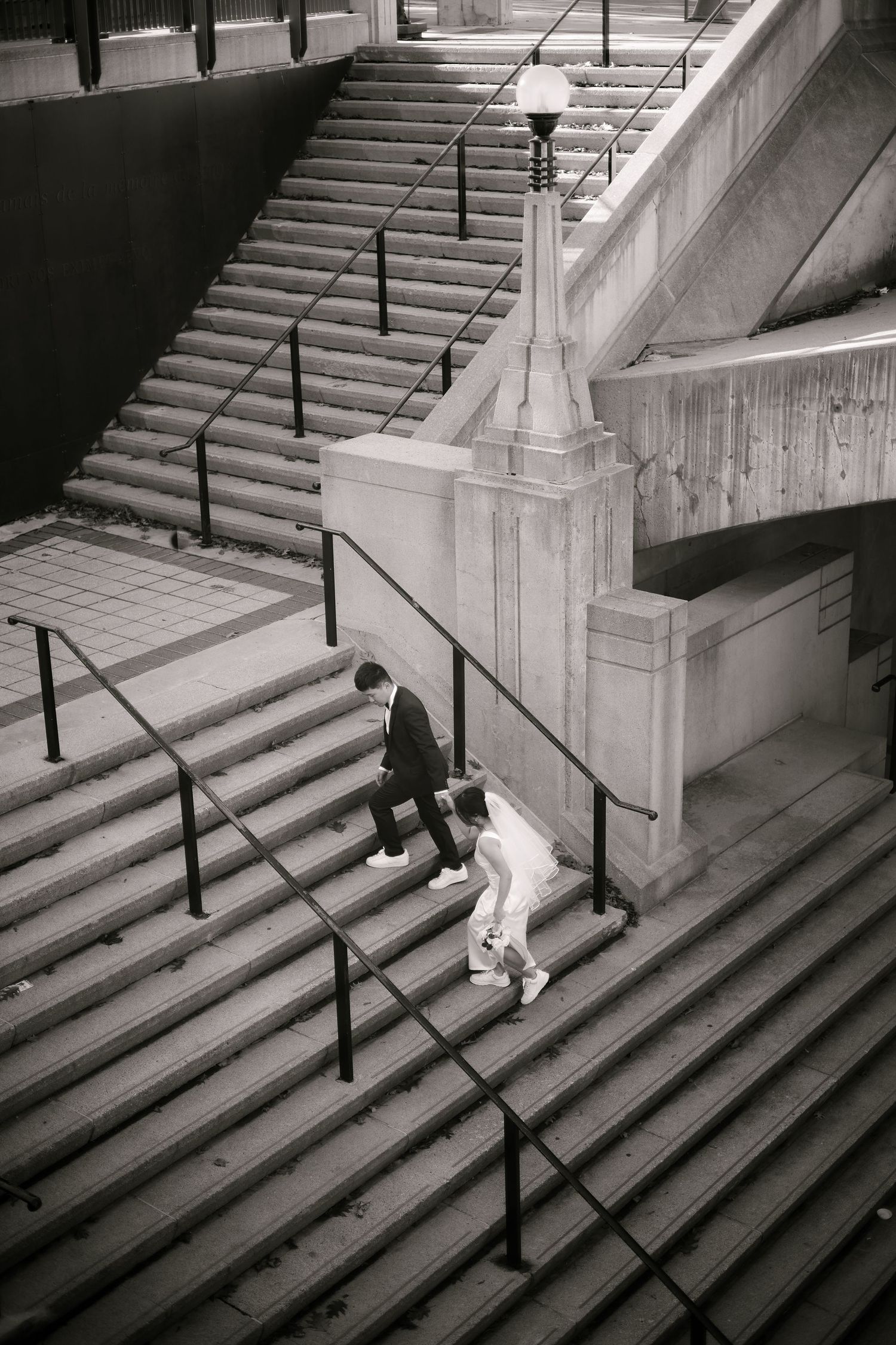 Black and white aerial view of two people descending wide urban stairs with metal railings and a lamppost.