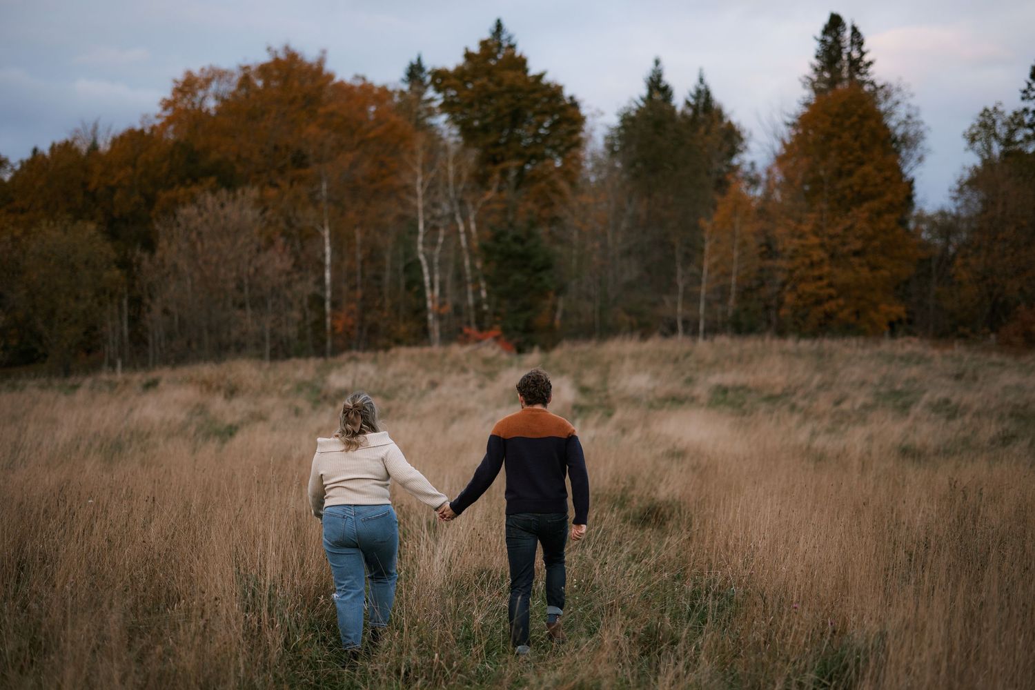 Couple holding hands walking through autumn field with colorful fall trees in background.
