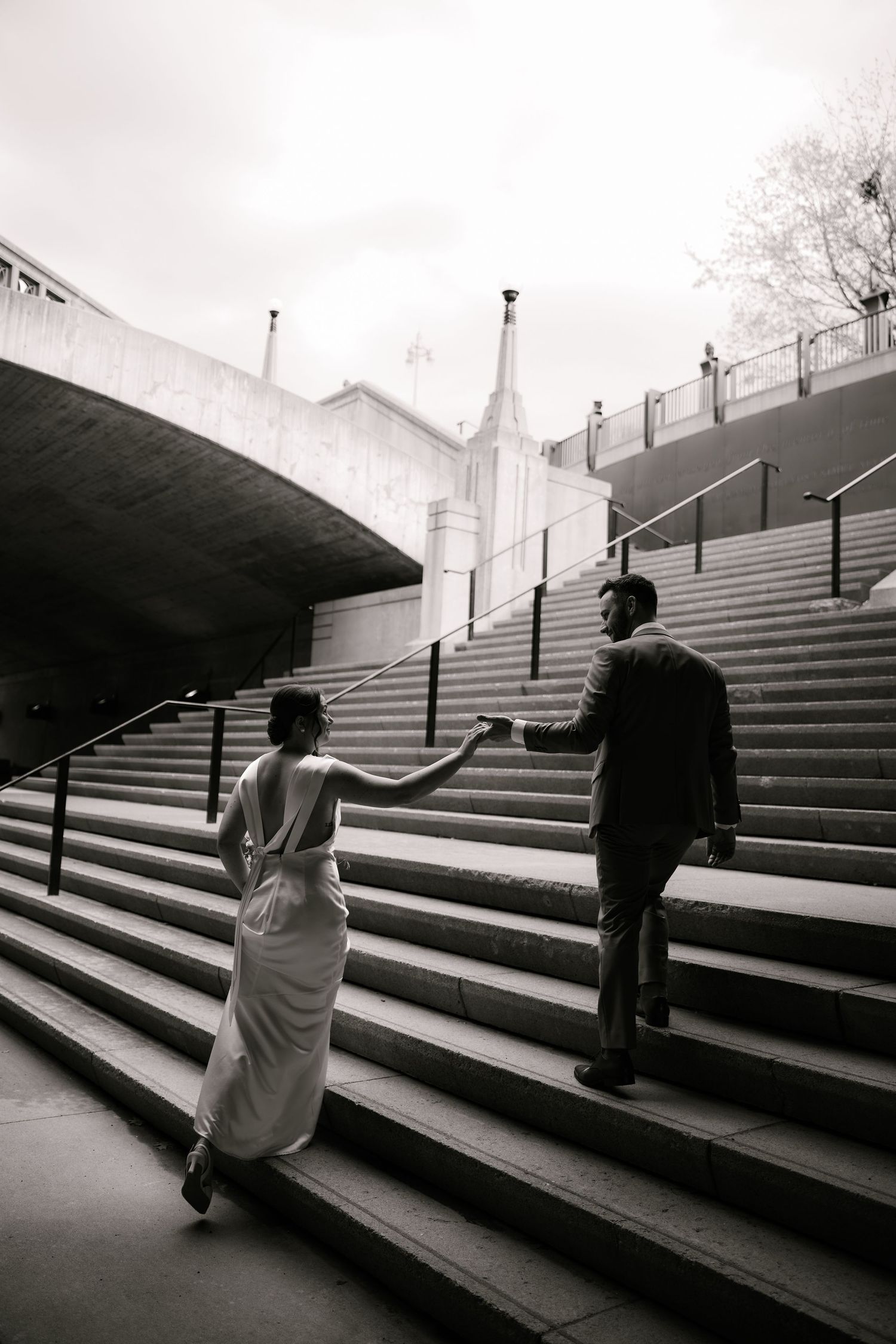 Black and white photo of a bride and groom holding hands while ascending grand outdoor stairs at their wedding.