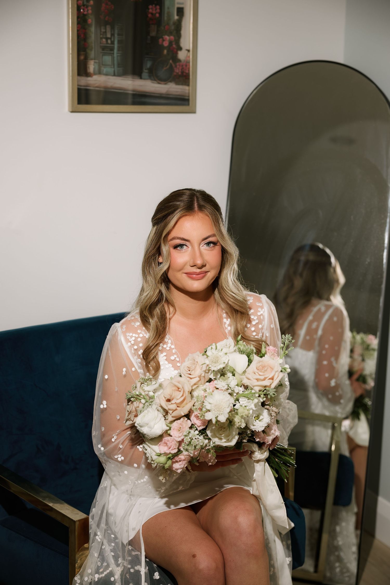 Bride in white lace robe holding blush and white floral bouquet, seated near arched mirror.