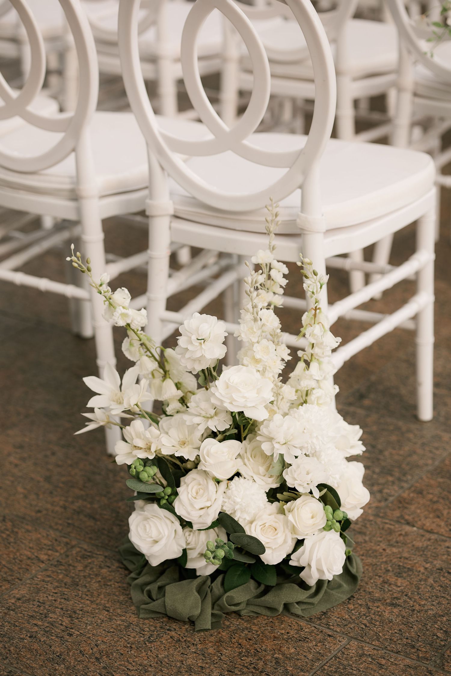 White floral arrangements with roses and greenery decorate white ceremony chairs.