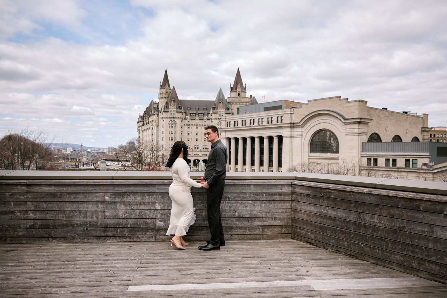 Couple holding hands on a wooden deck with Château Laurier and Canadian Museum of History in Ottawa background.