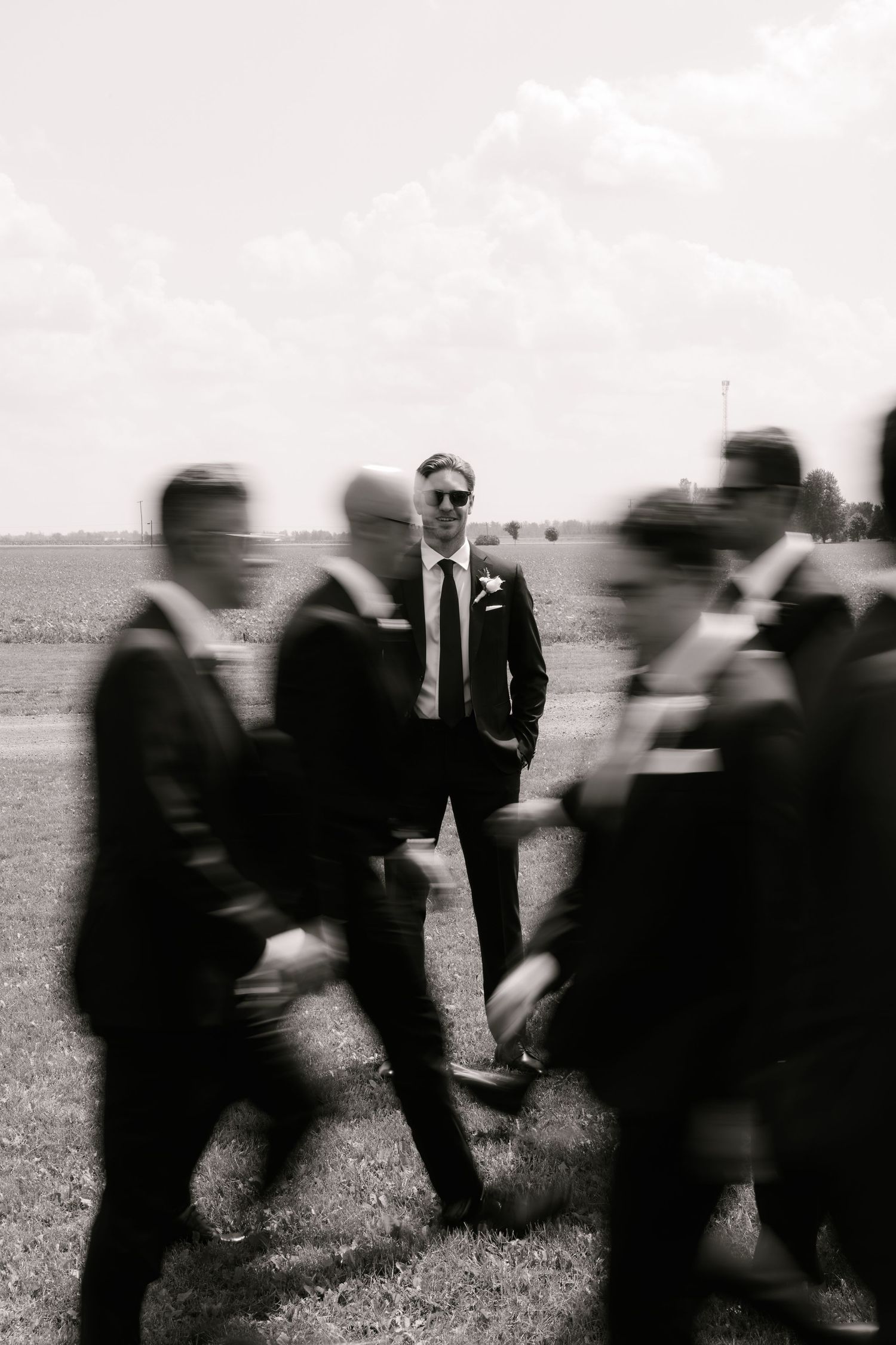 Black and white photo of groom standing still among blurred moving groomsmen in suits outdoors.
