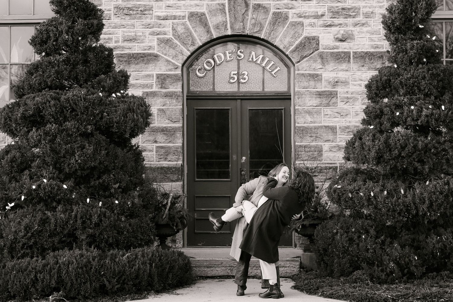 Couple embracing in front of Code's Mill stone building entrance, black and white engagement photo.