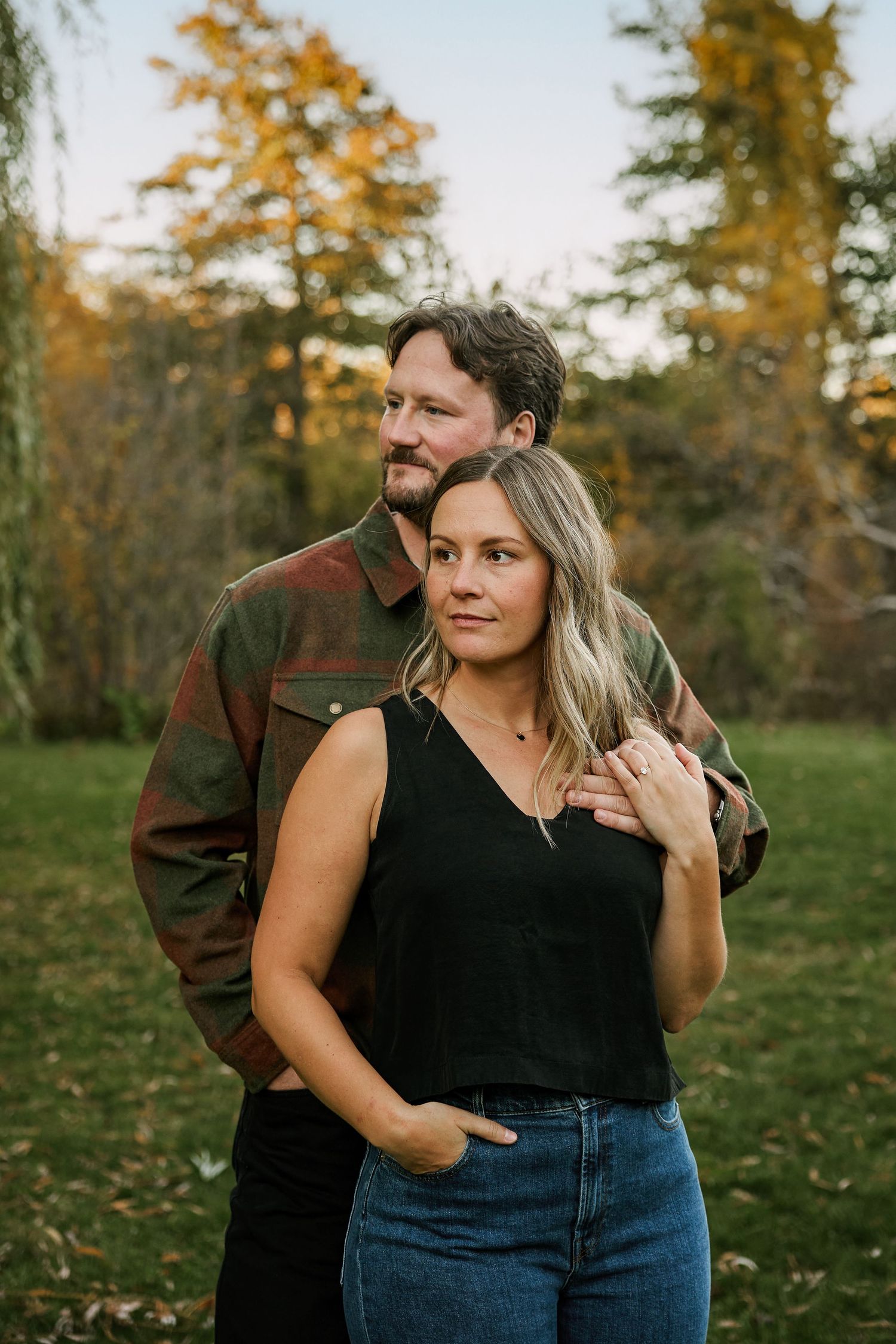 Couple embracing outdoors in autumn park, woman in black top and jeans, man in plaid jacket, fall foliage background.