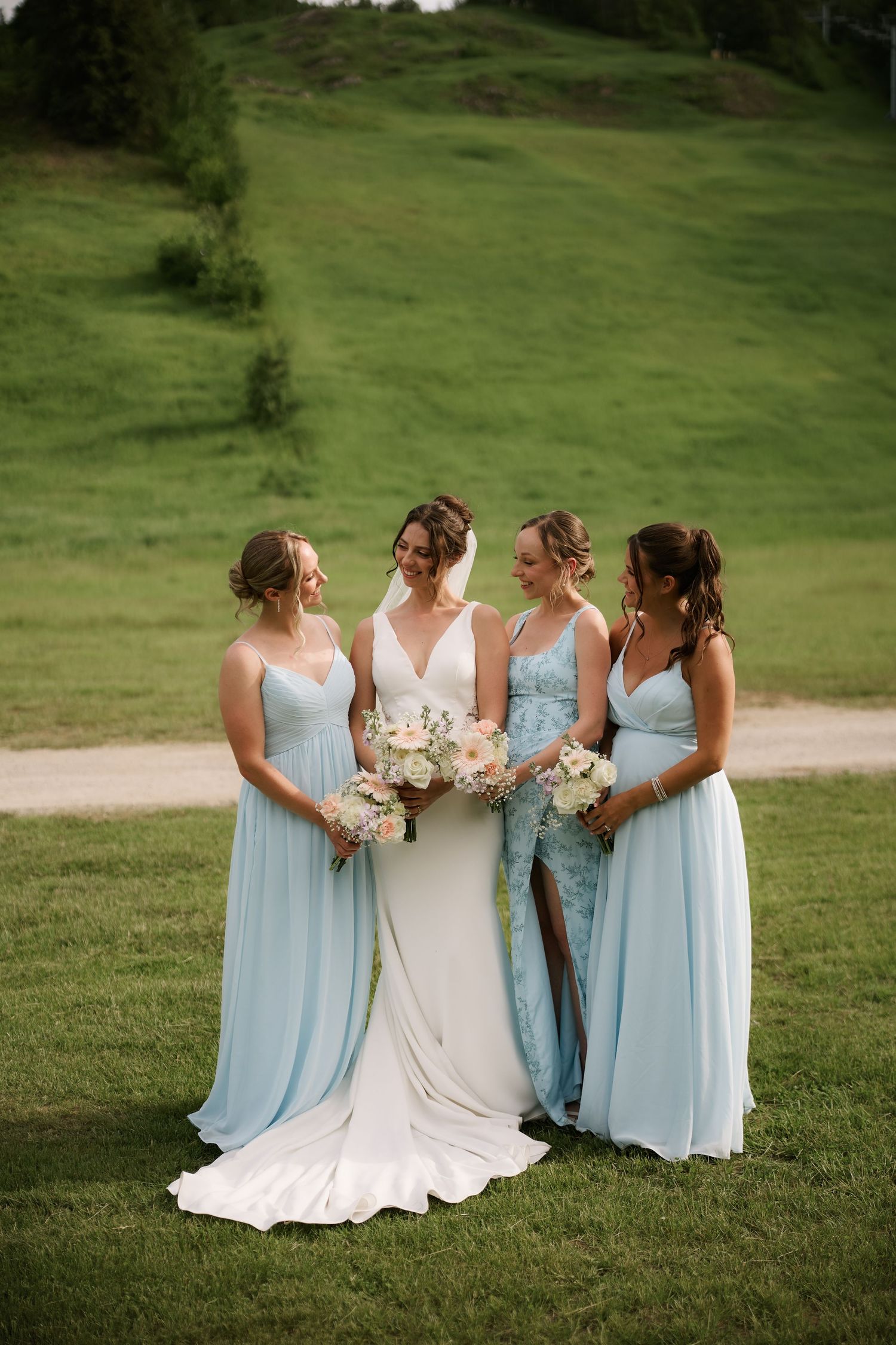 Bride in white gown with three bridesmaids in light blue dresses holding bouquets on green lawn.
