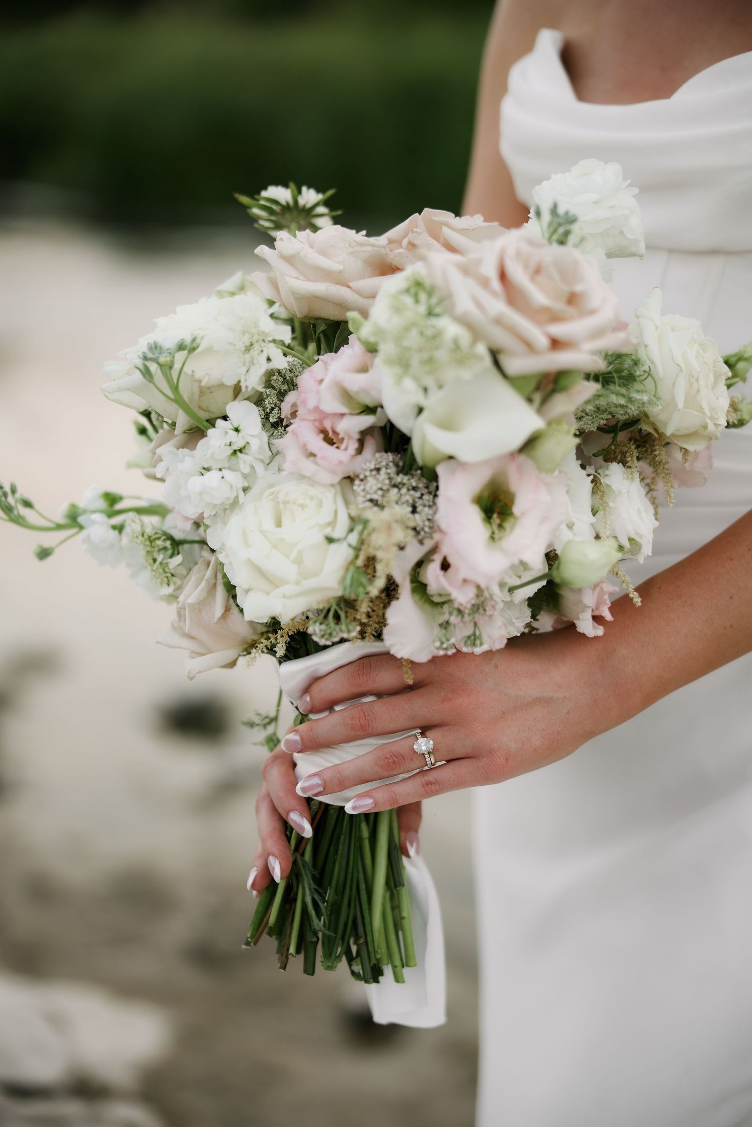 Bride holding romantic bouquet of blush roses, white ranunculus, and greenery with diamond engagement ring visible.