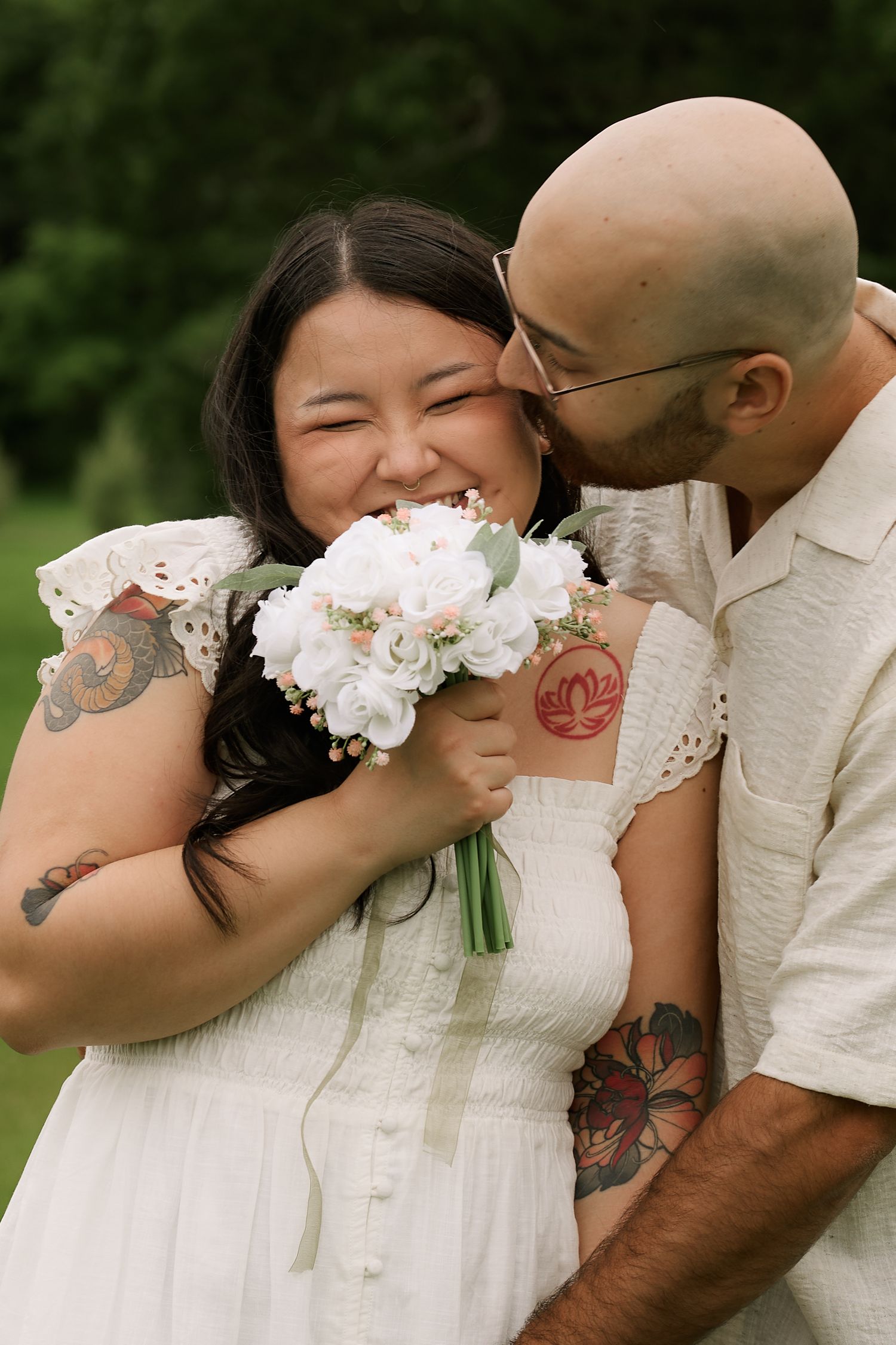 Tattooed couple sharing a romantic moment, bride in white dress holding bouquet, groom kissing her cheek outdoors.
