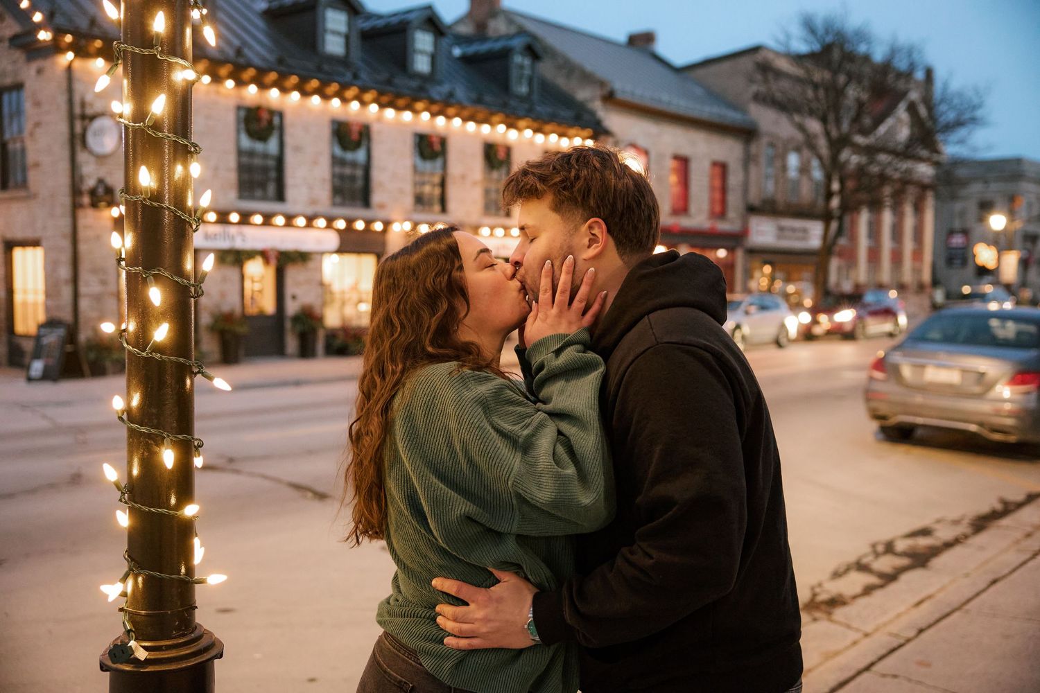 Couple sharing a romantic kiss by a festive light-wrapped lamppost on a charming small town street at dusk.