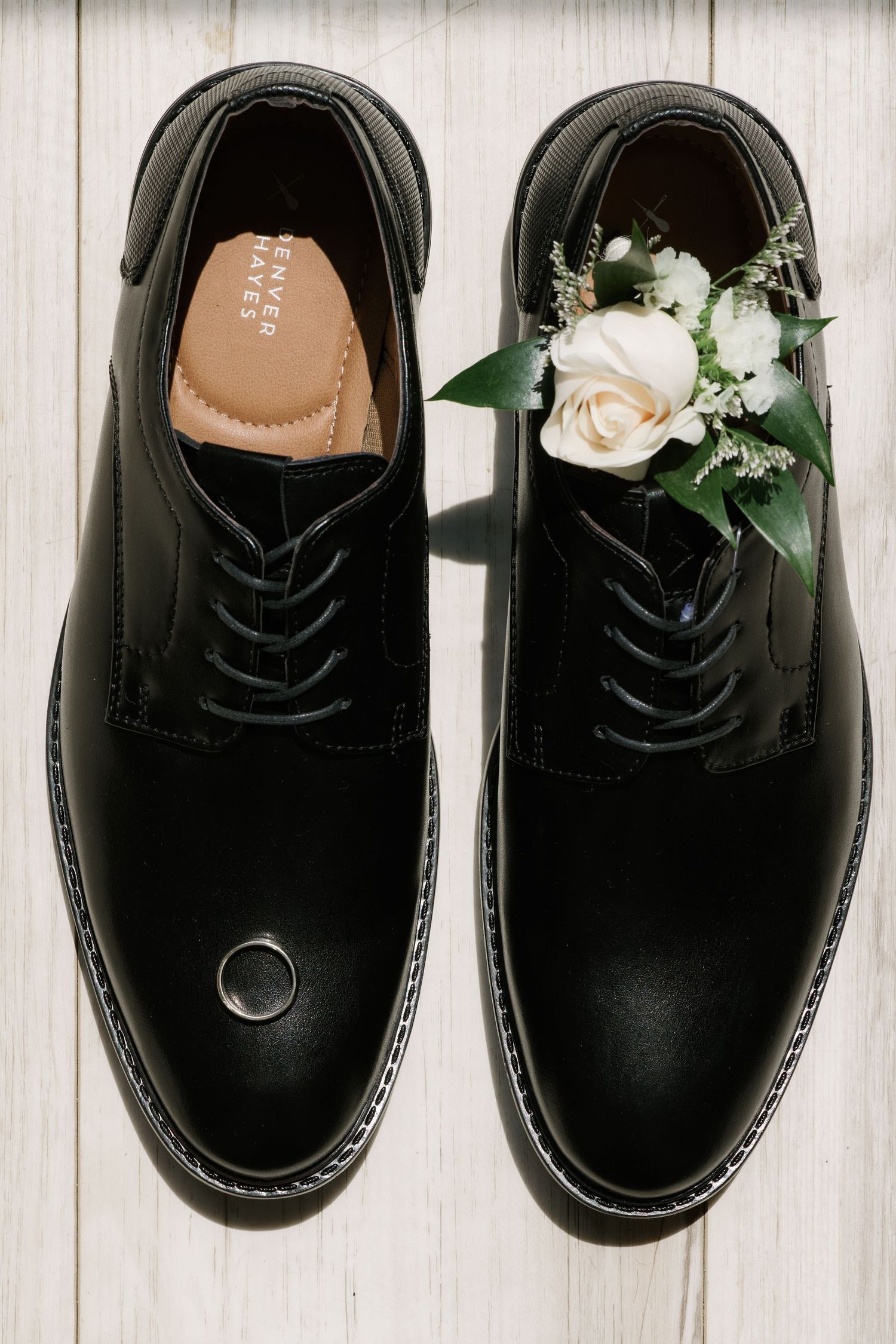 Black dress shoes with a white rose boutonniere and wedding ring, arranged on a white wooden surface.