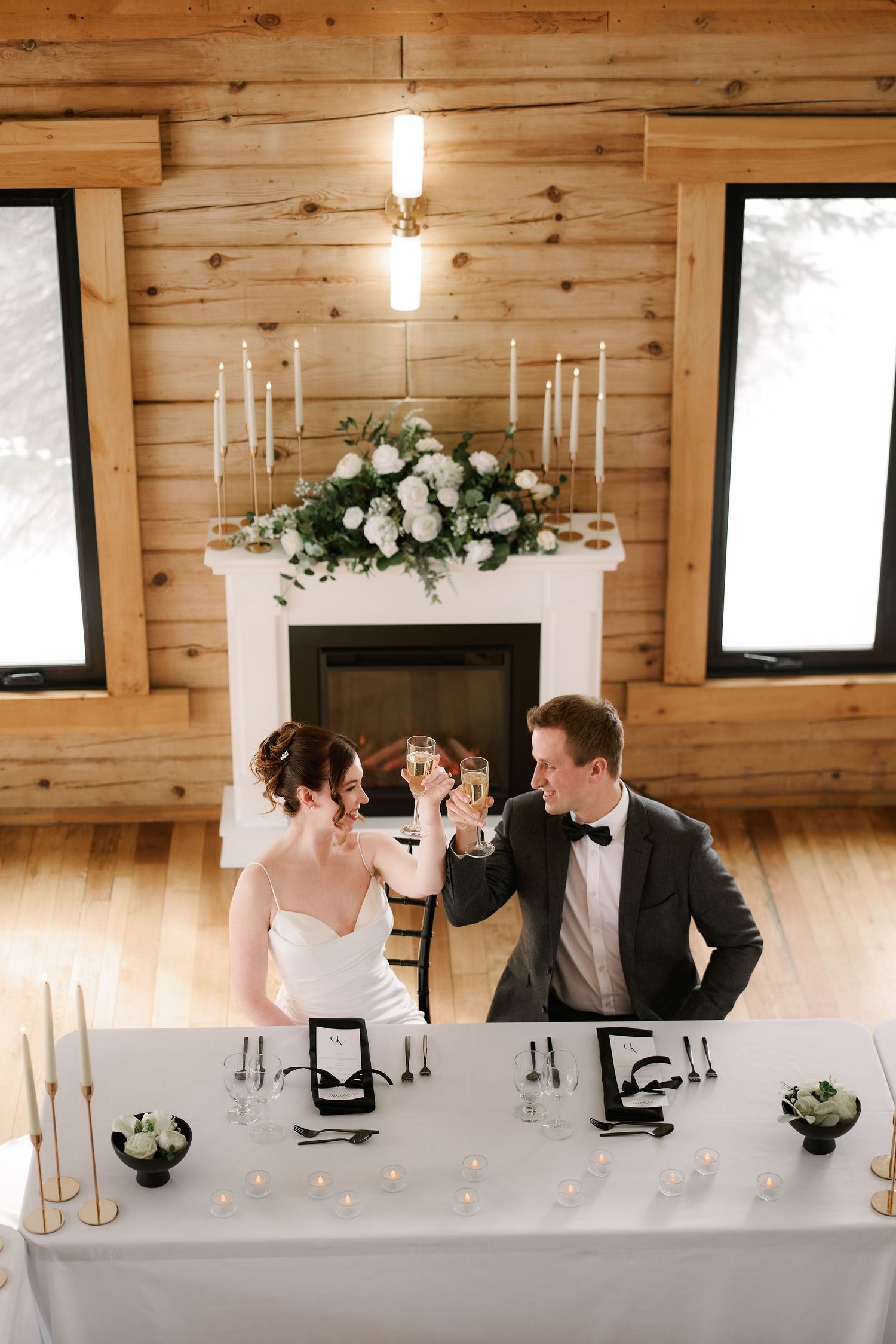 Newlyweds sharing a drink at an elegant table with floral decor and candles in a rustic wooden venue.