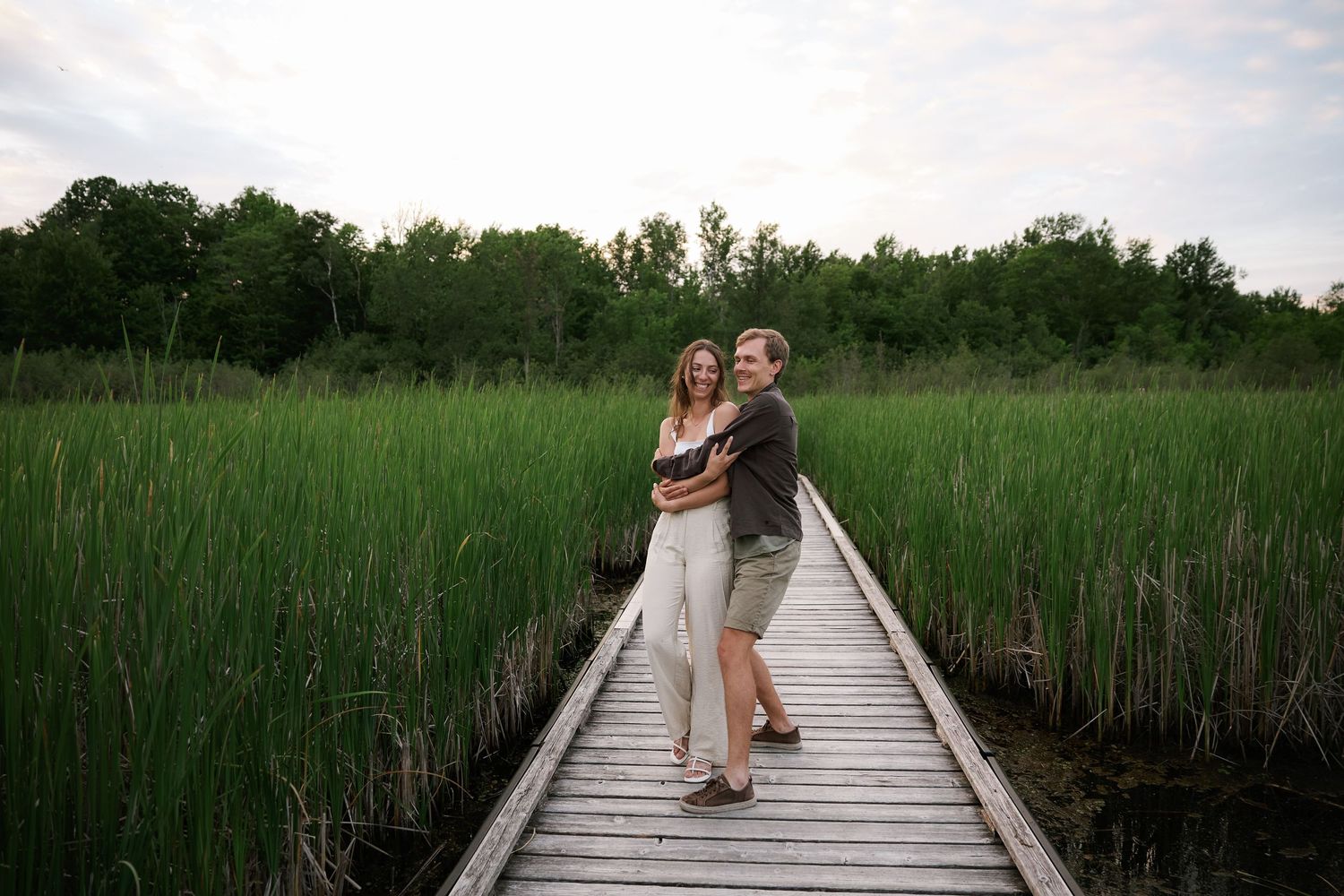 A romantic couple sits together on a wooden boardwalk path through tall marsh grasses.
