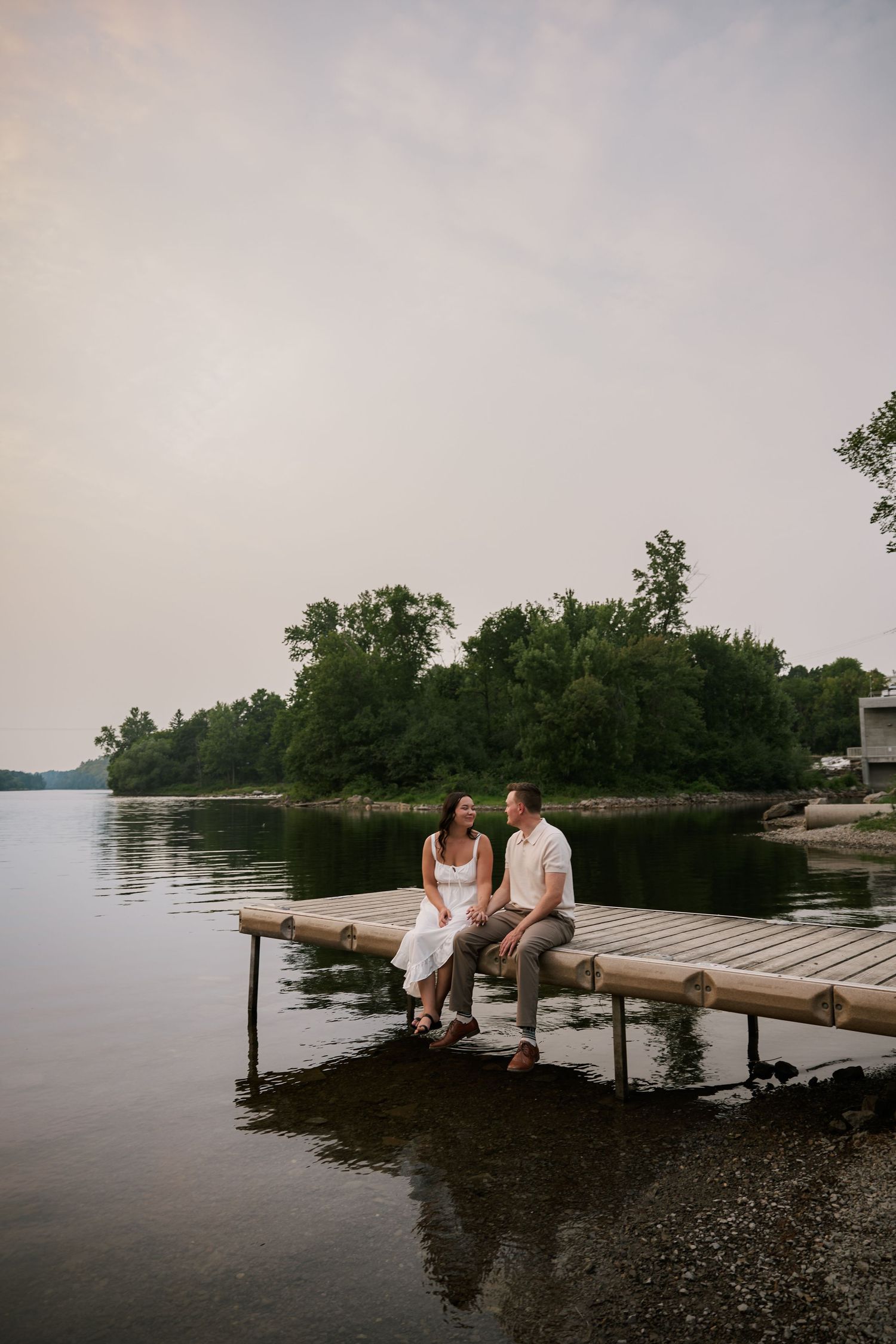 A couple sits together on a wooden dock overlooking a tranquil lake at sunset.