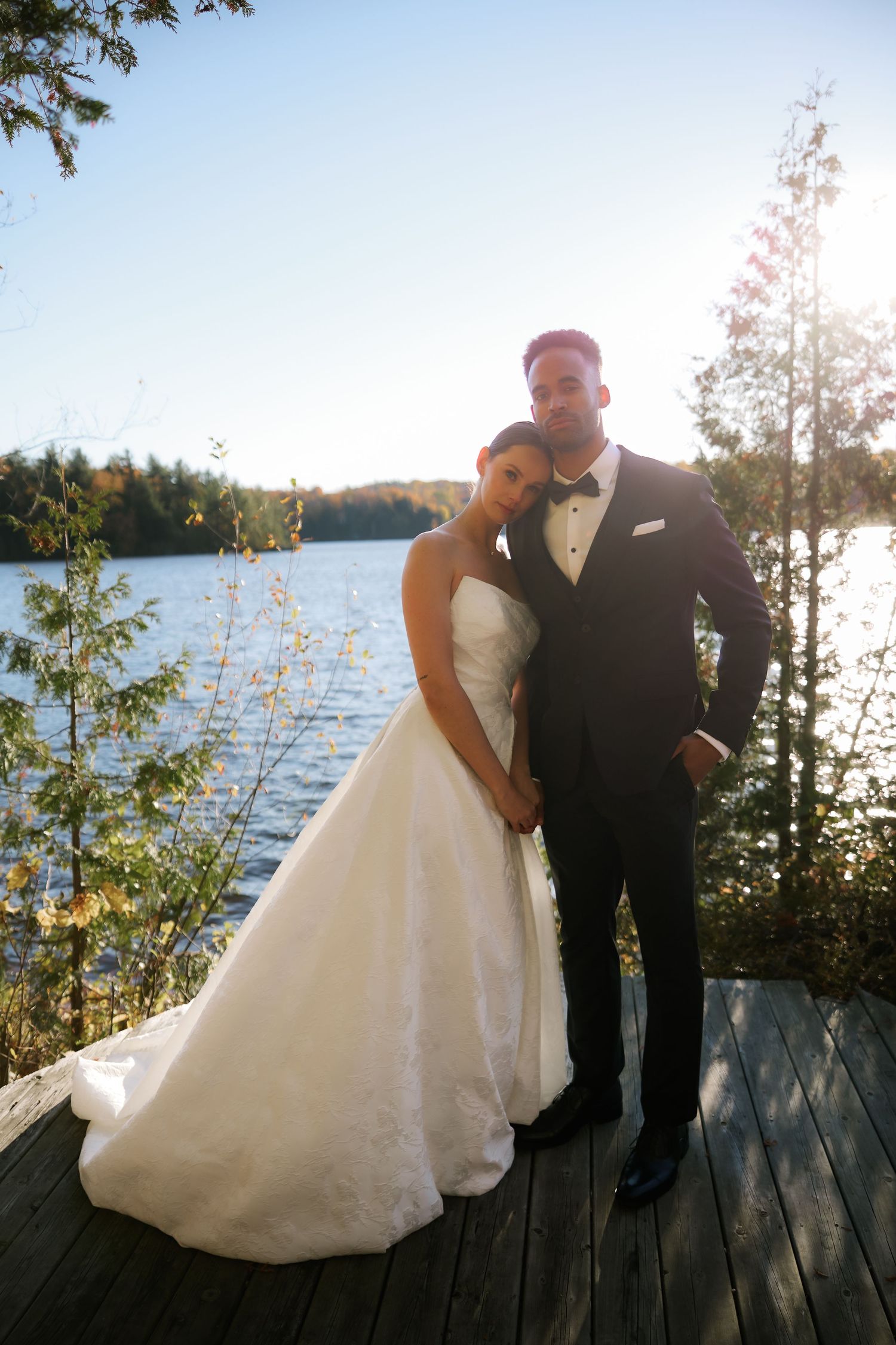 Bride in a strapless white gown rests her head on her groom's chest on a wooden lakeside dock, with trees and sunlit water behind.