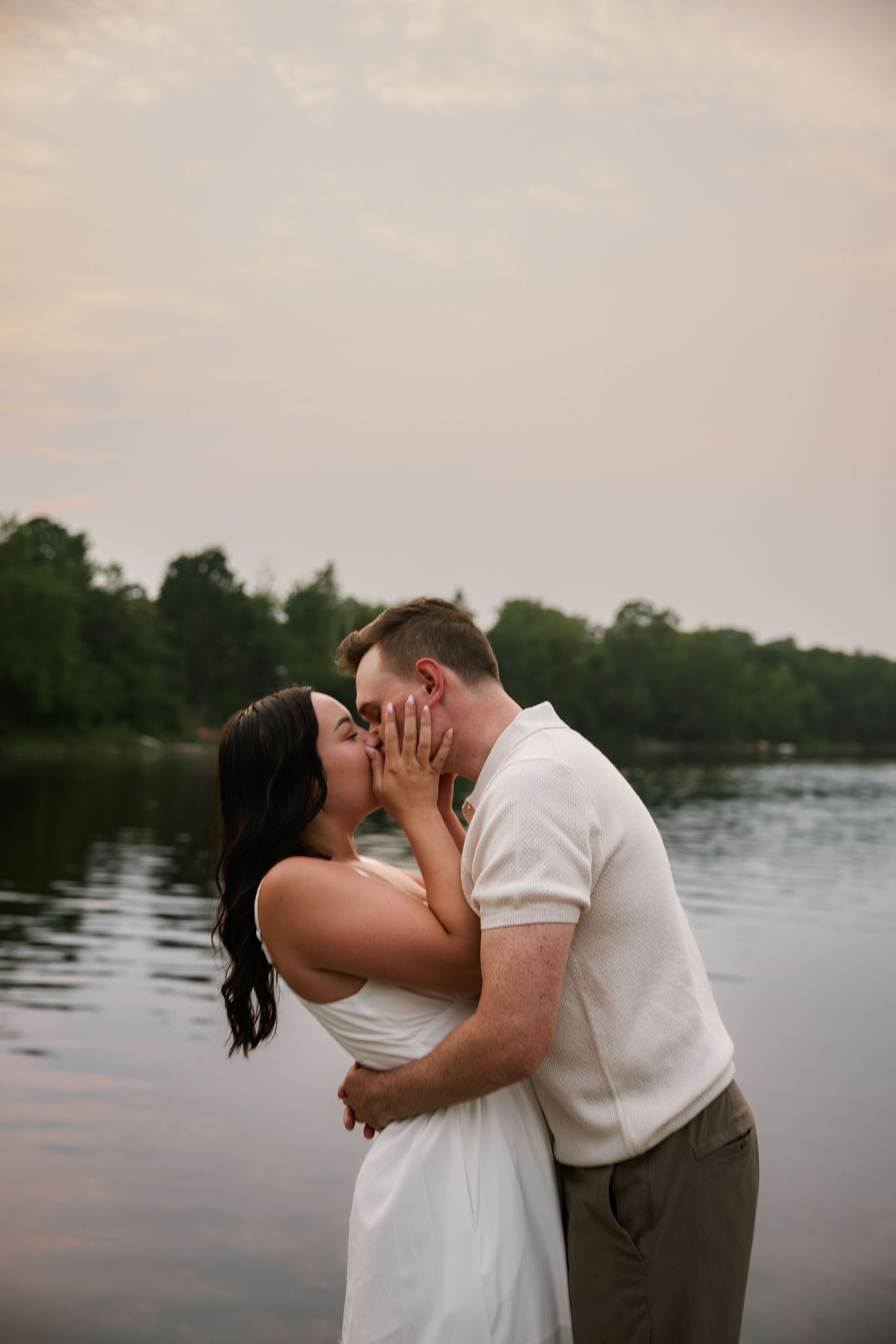 Couple sharing a romantic kiss by a serene lake at sunset, woman in white dress, man in white shirt and khaki pants.
