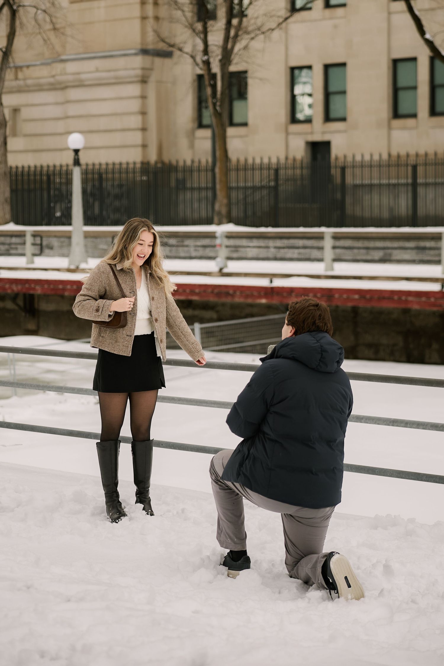 Person proposing on one knee in the snow to a surprised woman wearing a tan jacket and black skirt outdoors.