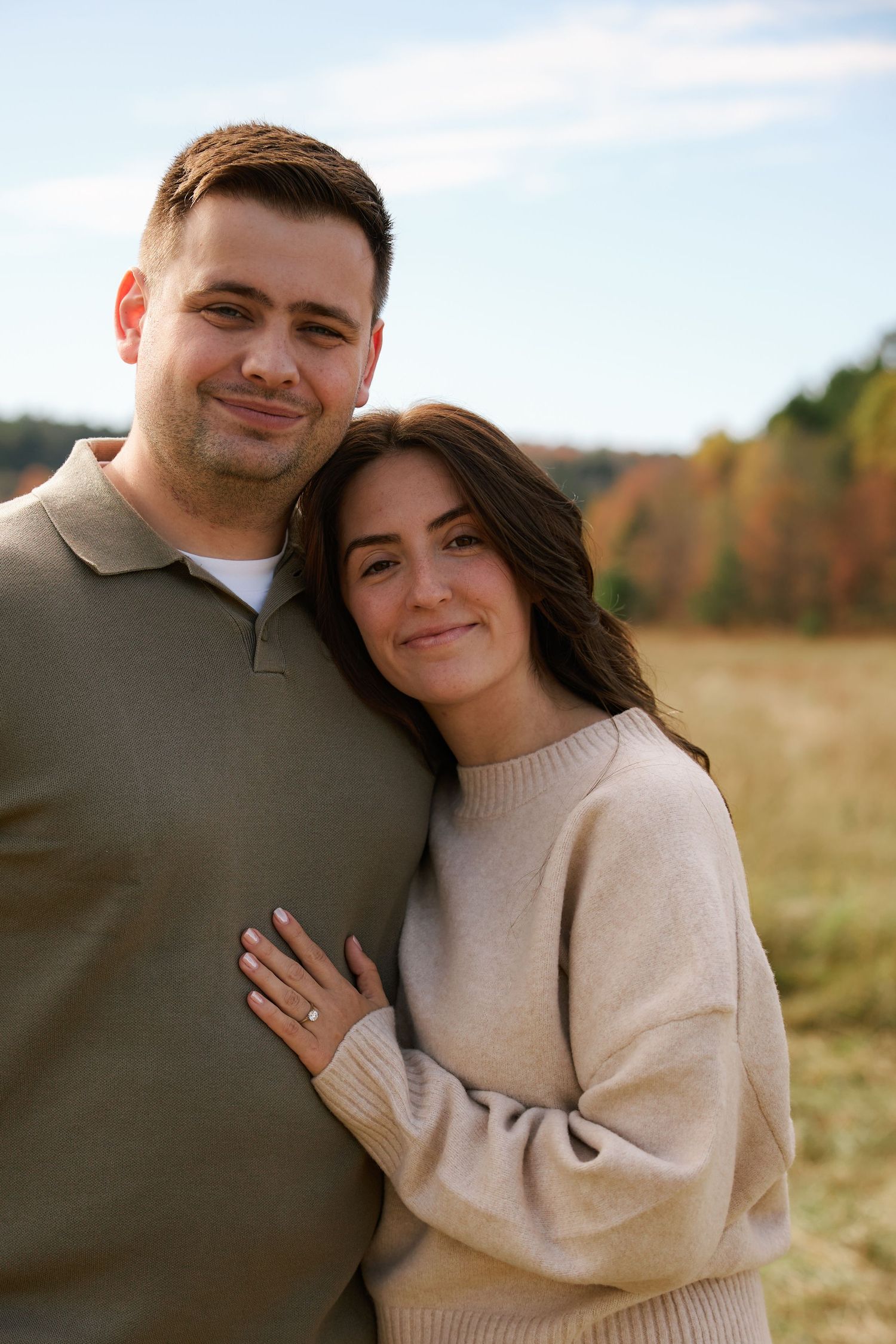 Couple smiling outdoors in autumn field, woman leaning on mans shoulder, wearing cozy sweaters, engagement ring visible.