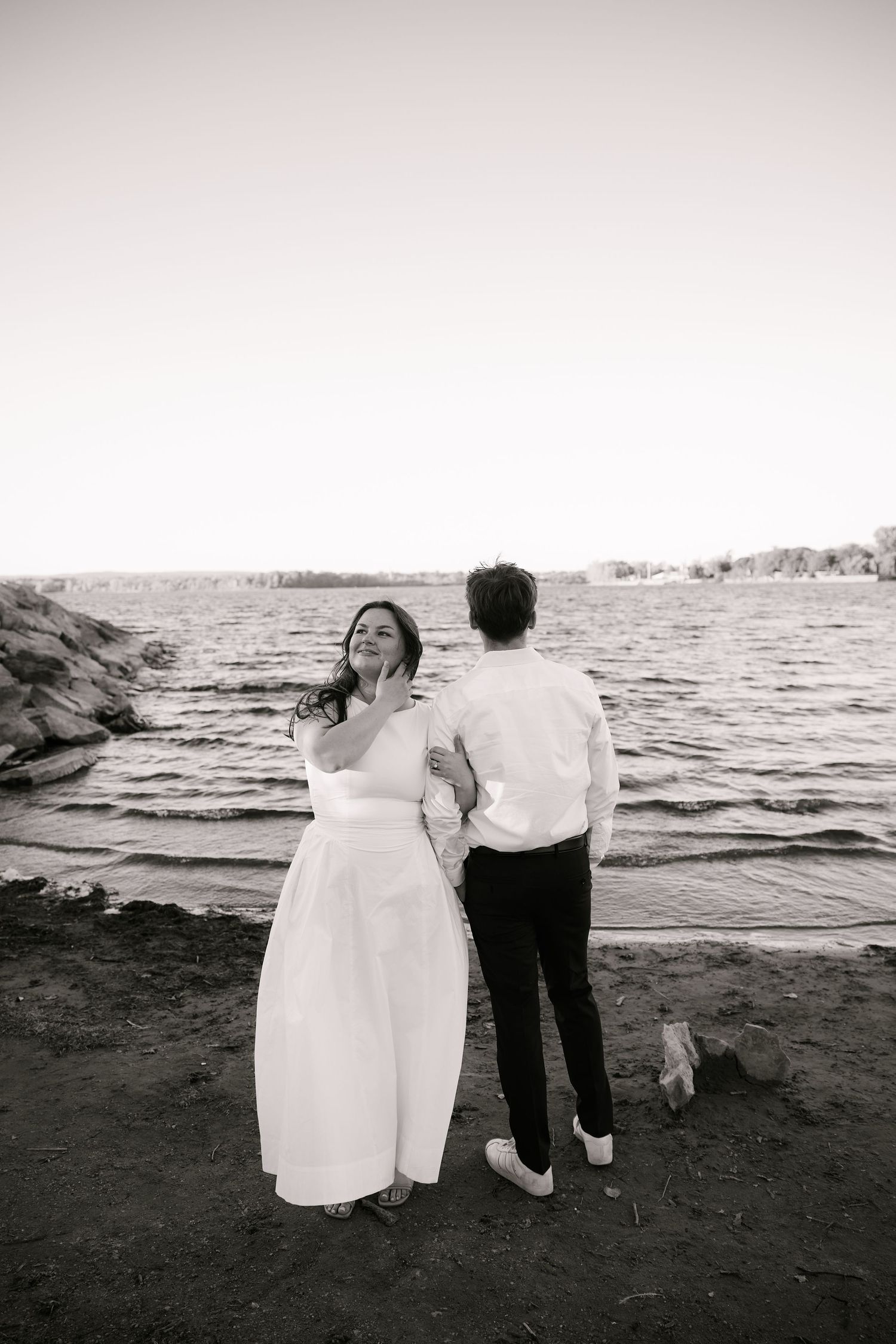 Black and white photo of a bride and groom standing by the water's edge, the bride laughing joyfully.