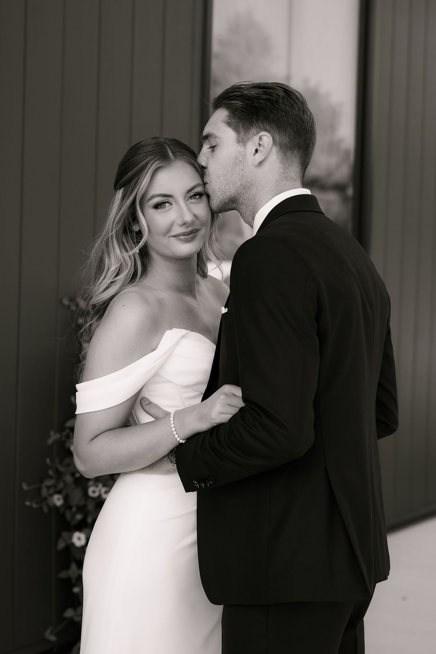 Black and white photo of a groom kissing his bride on the cheek as she smiles in an off-shoulder wedding gown.