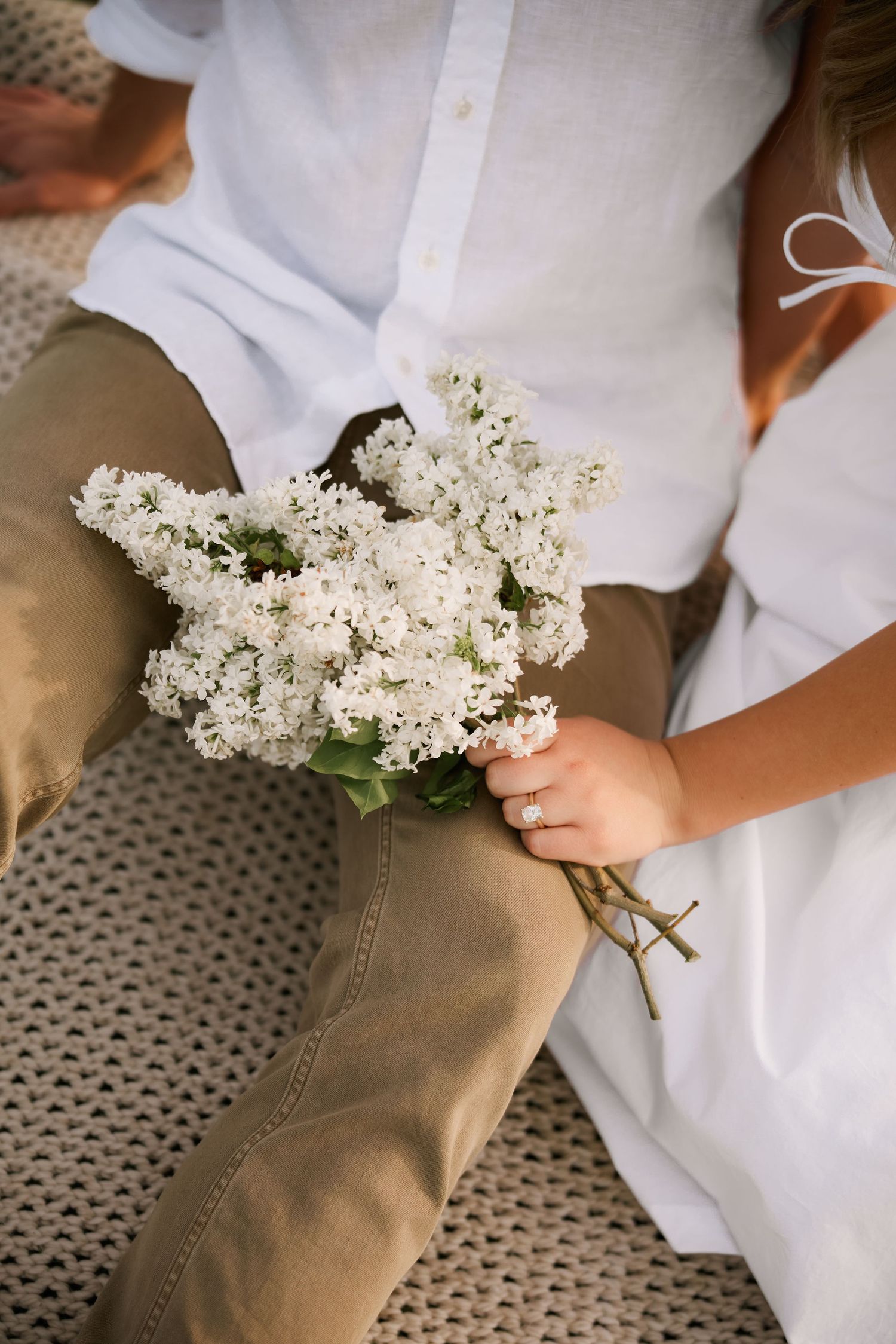 Person holding white lilac bouquet while sitting, wearing white linen shirt and khaki pants, with diamond engagement ring.