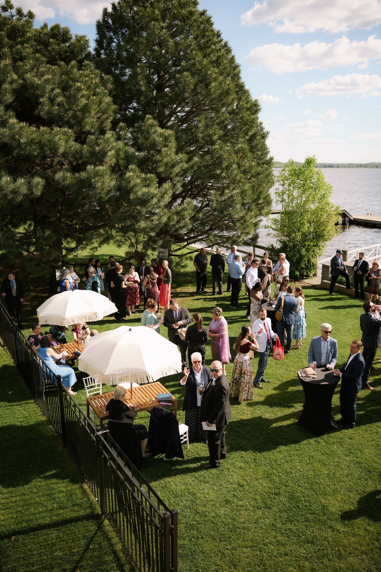 Outdoor garden party near a lake with guests mingling on green lawn under white umbrellas beside tall pine trees.
