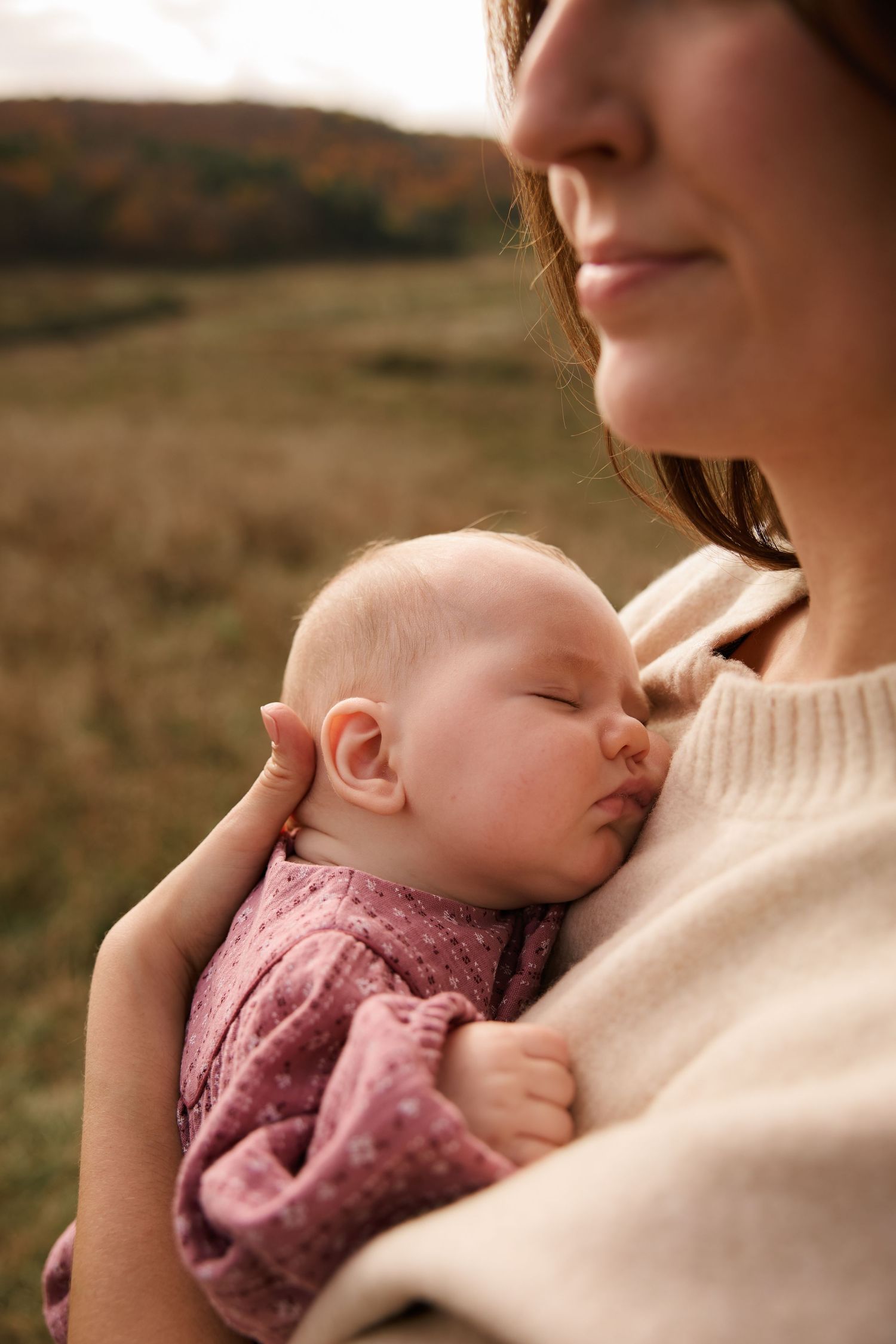 Baby in pink dress rests peacefully against cream sweater in outdoor setting.