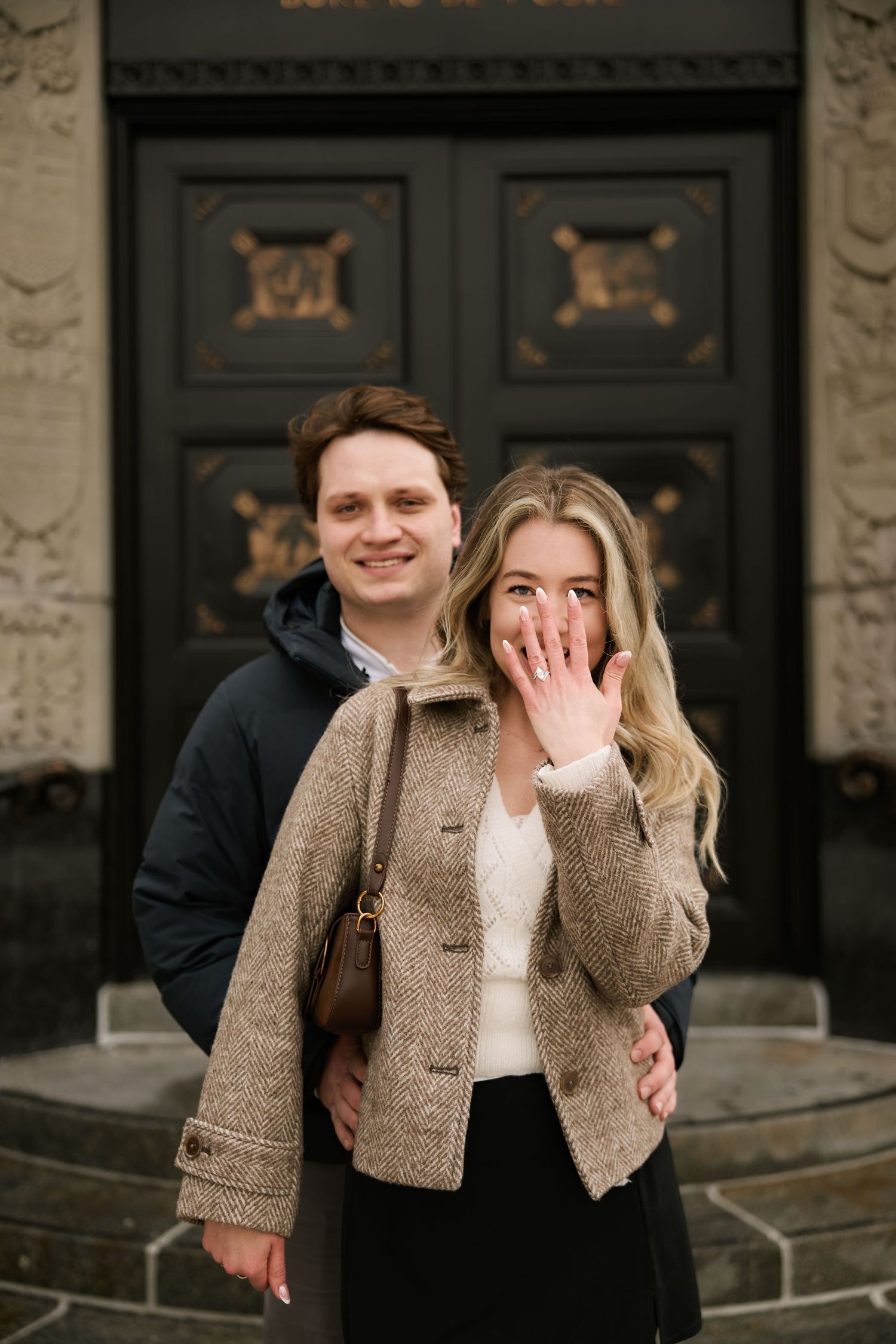 A couple laughs together while standing in front of dark wooden doors on stone steps.