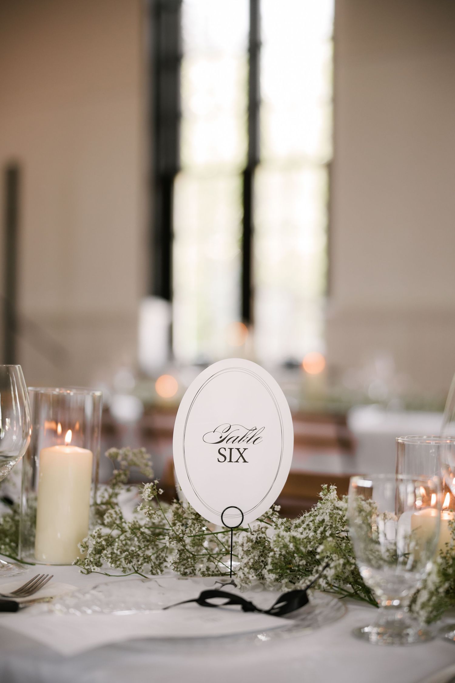 Elegant wedding table six sign with white oval frame, candles, and baby's breath floral centerpiece on white linen.