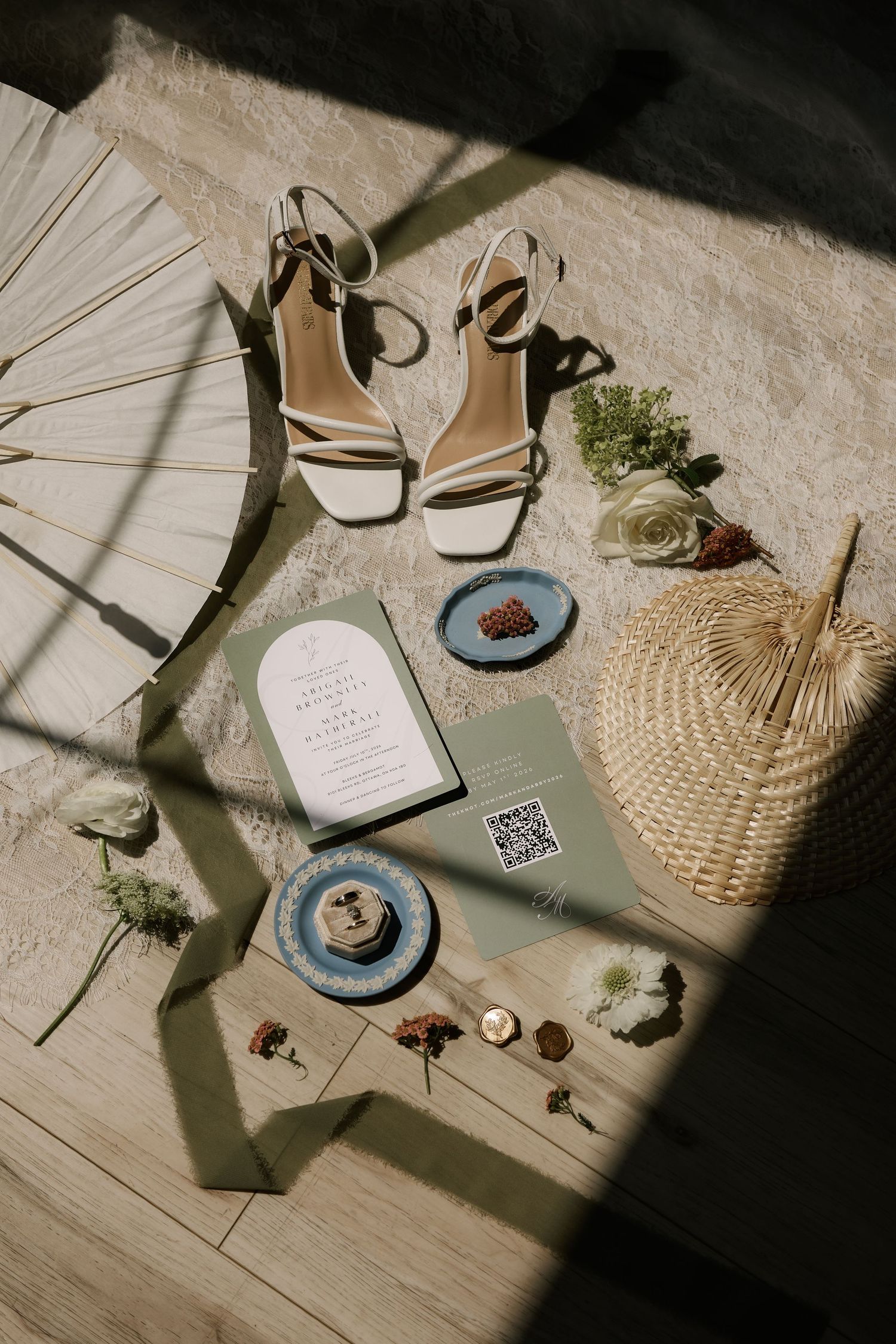 Flat lay of bridal accessories including white heeled sandals, wedding invitation, flowers, and woven basket in natural light.