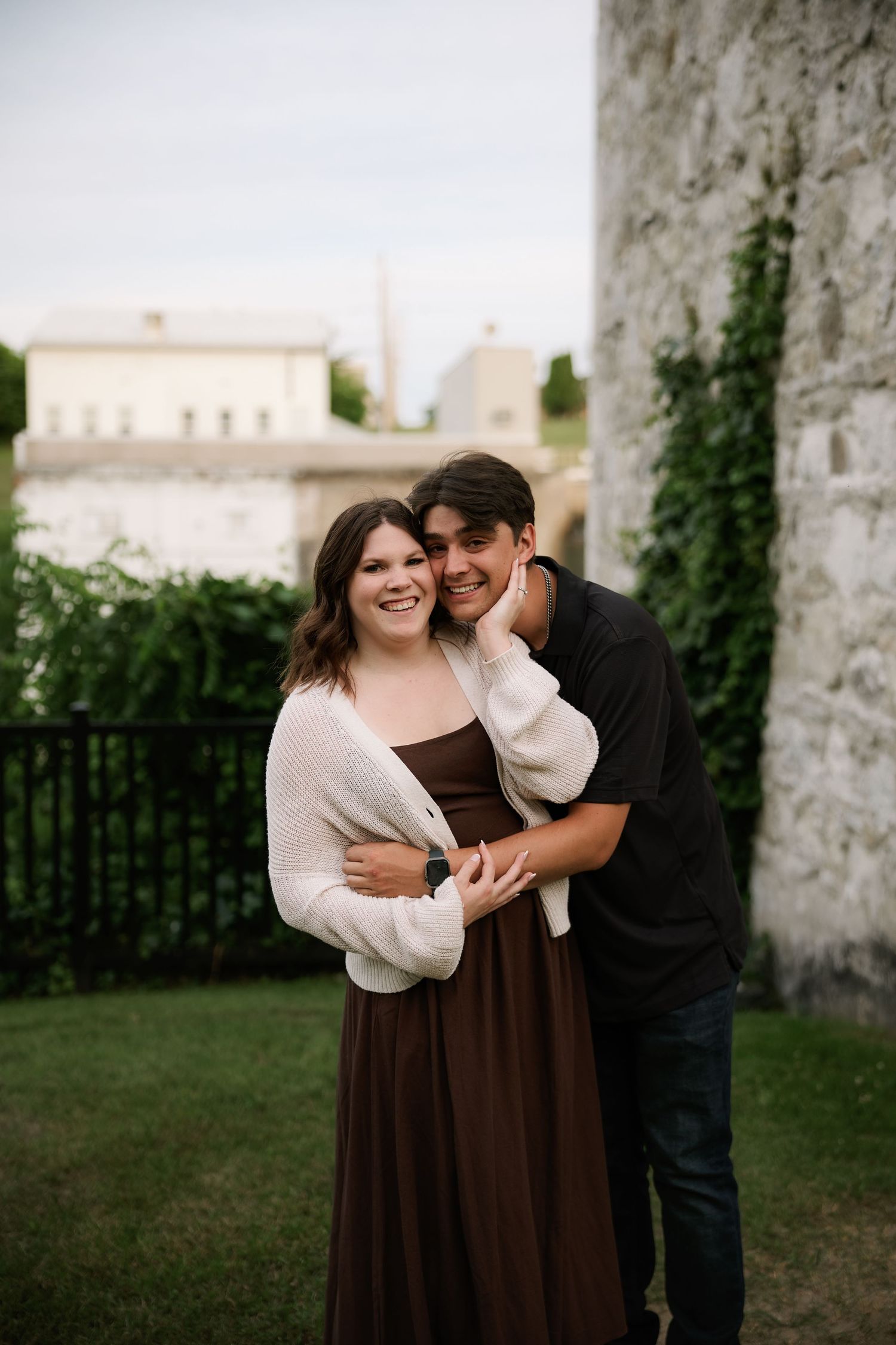 A smiling couple poses outdoors near a stone wall, the man embracing the woman in a brown dress and white cardigan.