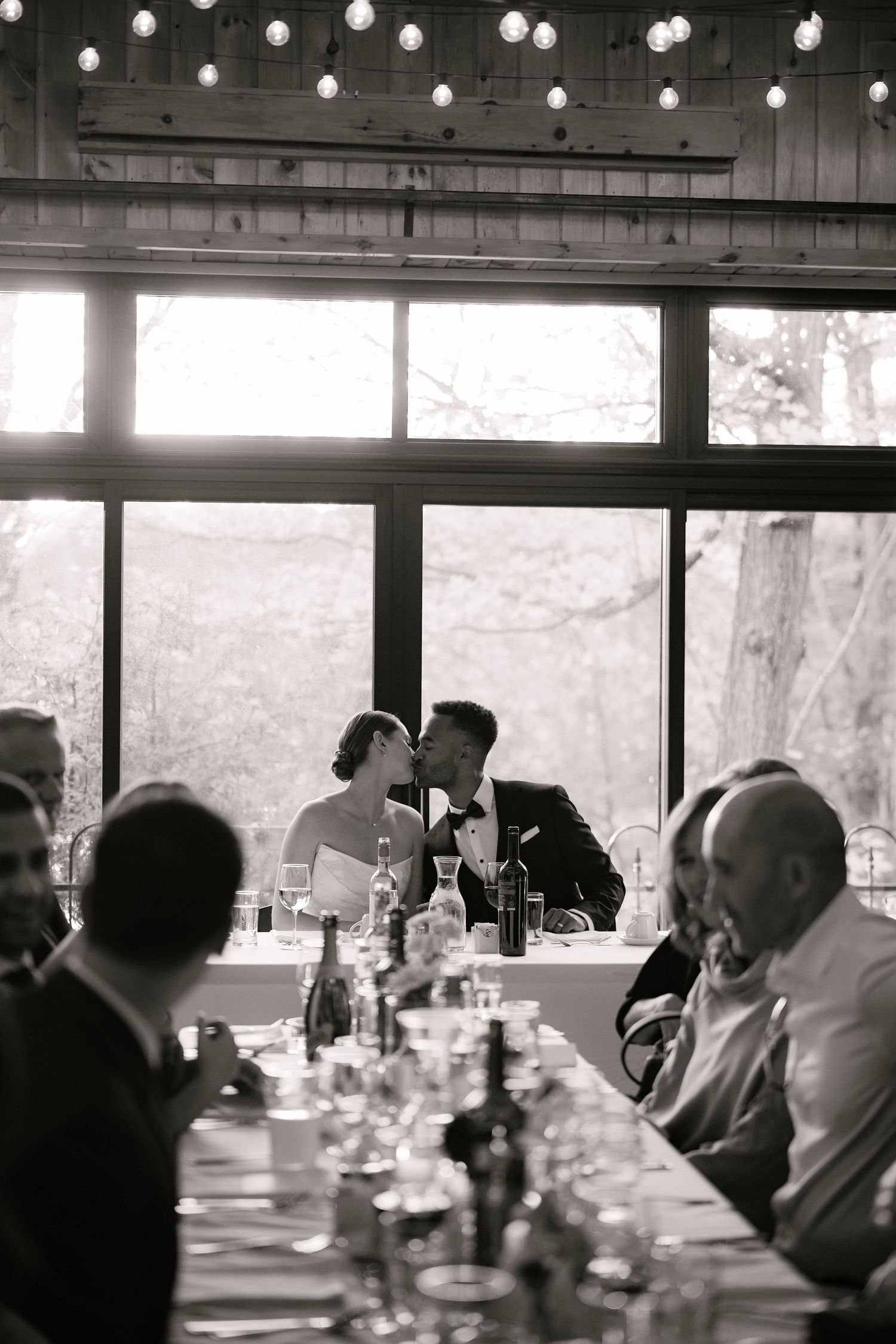 Black and white photo of a bride and groom kissing at a long reception table with string lights and large windows.