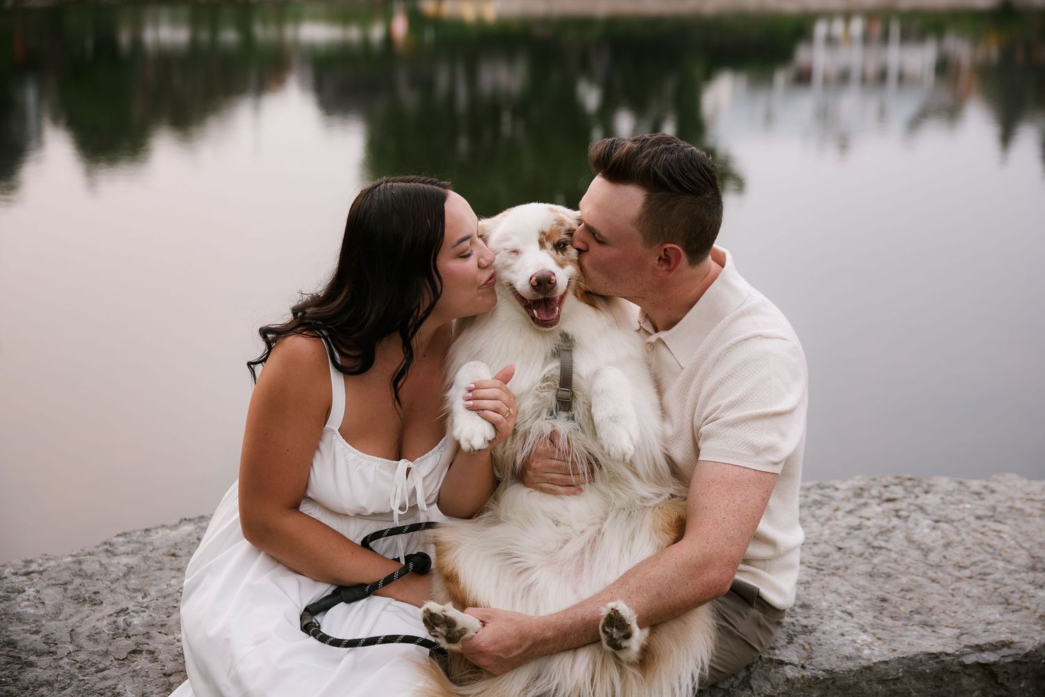 A couple shares a tender moment with their white dog by a peaceful lake.