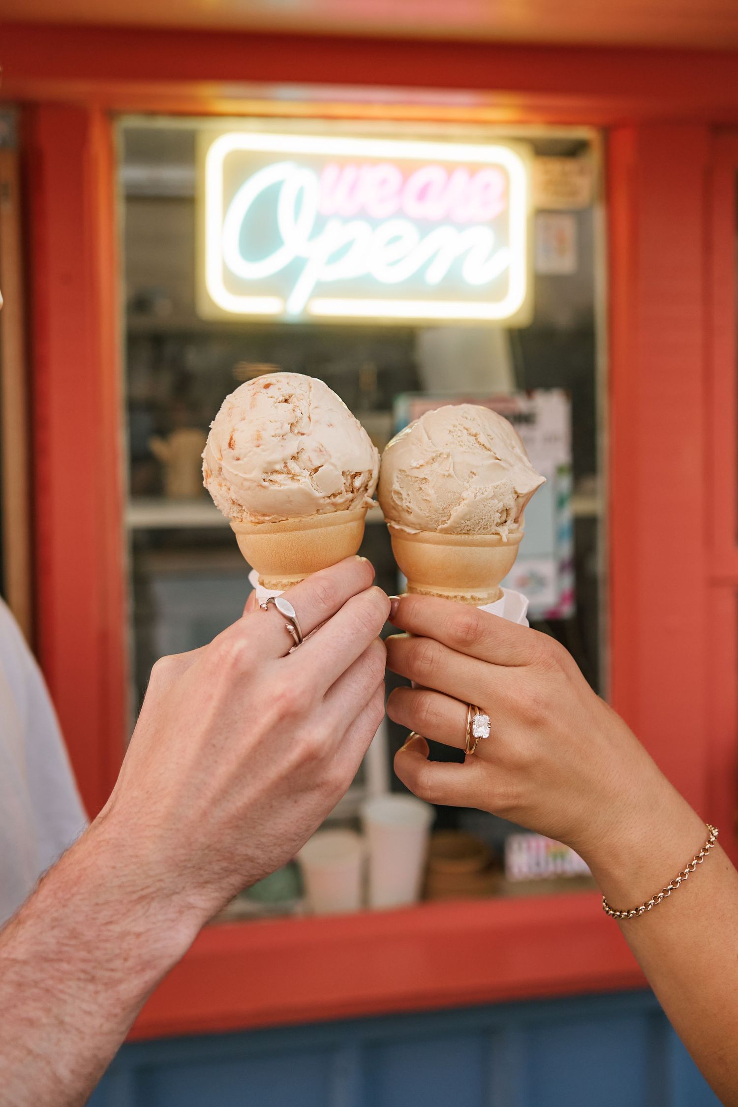 Two hands holding ice cream cones in front of an illuminated shop sign.