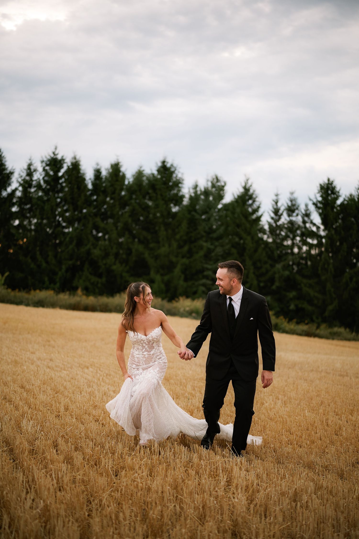 A couple walks hand in hand through a golden wheat field with evergreen trees in the background on their wedding day.