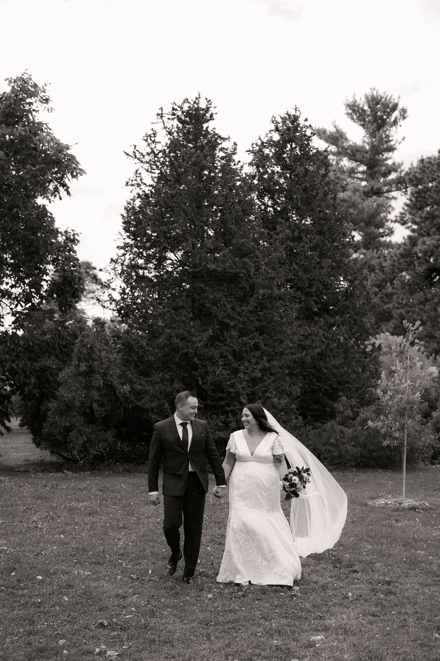 A couple in formal wedding attire walks together across a grassy field in black and white photography.