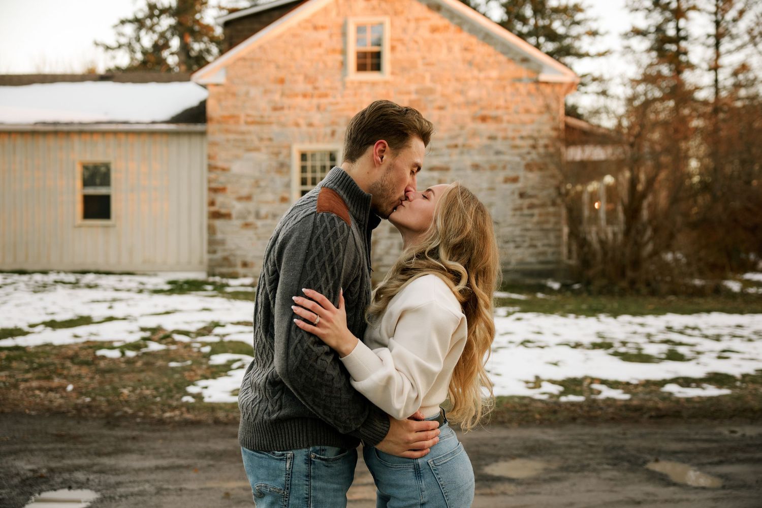 Couple sharing a romantic kiss outdoors in winter, embracing near a rustic stone building with snow on the ground.