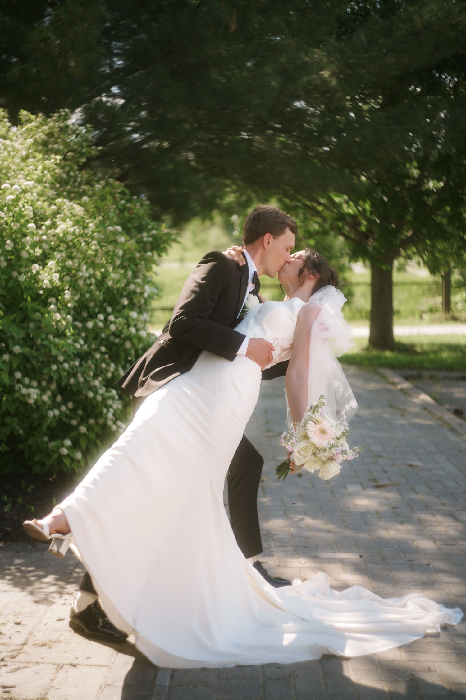 Groom dips bride for a kiss on a tree-lined path, bride holding a pastel bouquet in a white gown.