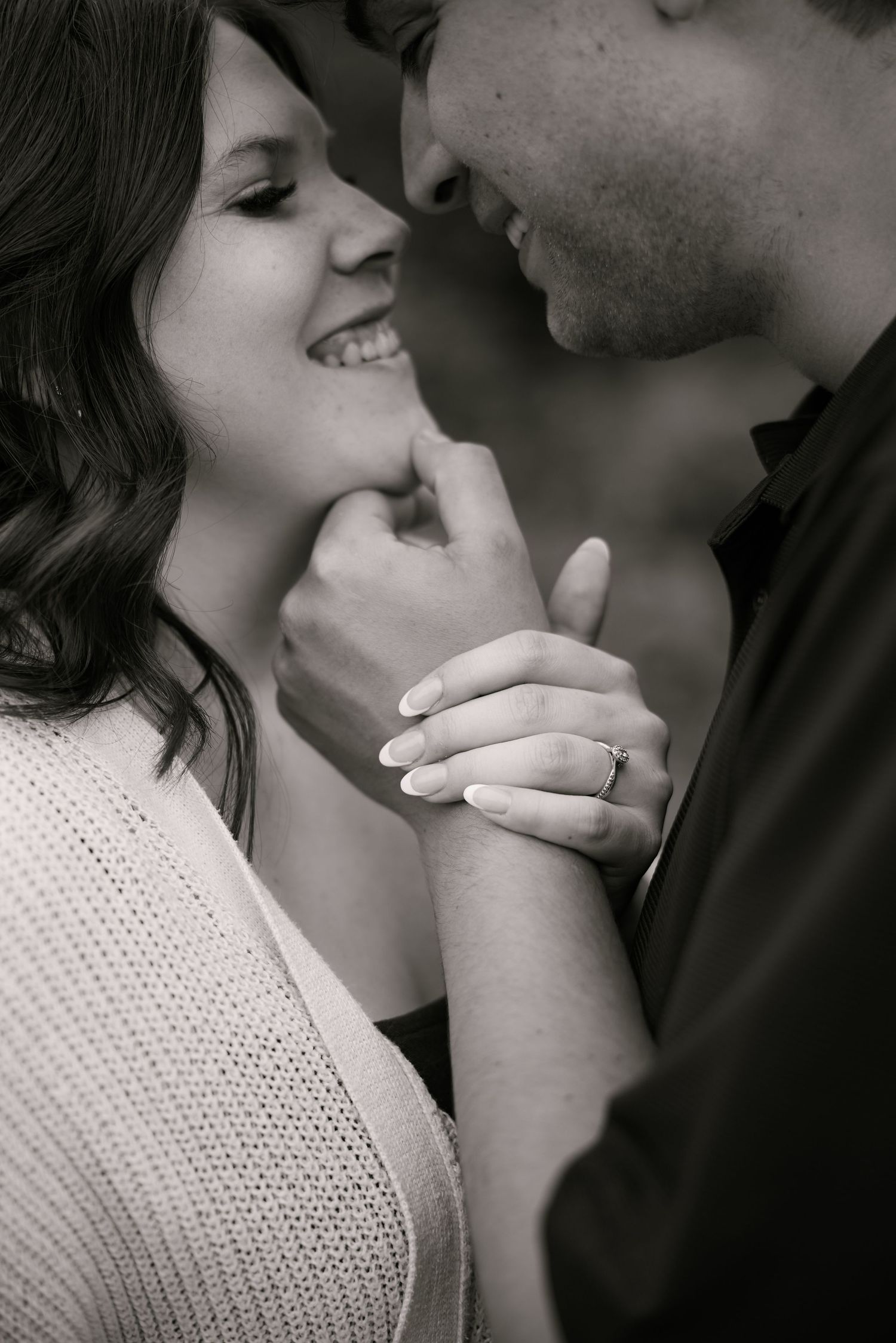 Black and white photo of a smiling couple holding hands, woman wearing an engagement ring, sharing a romantic moment.