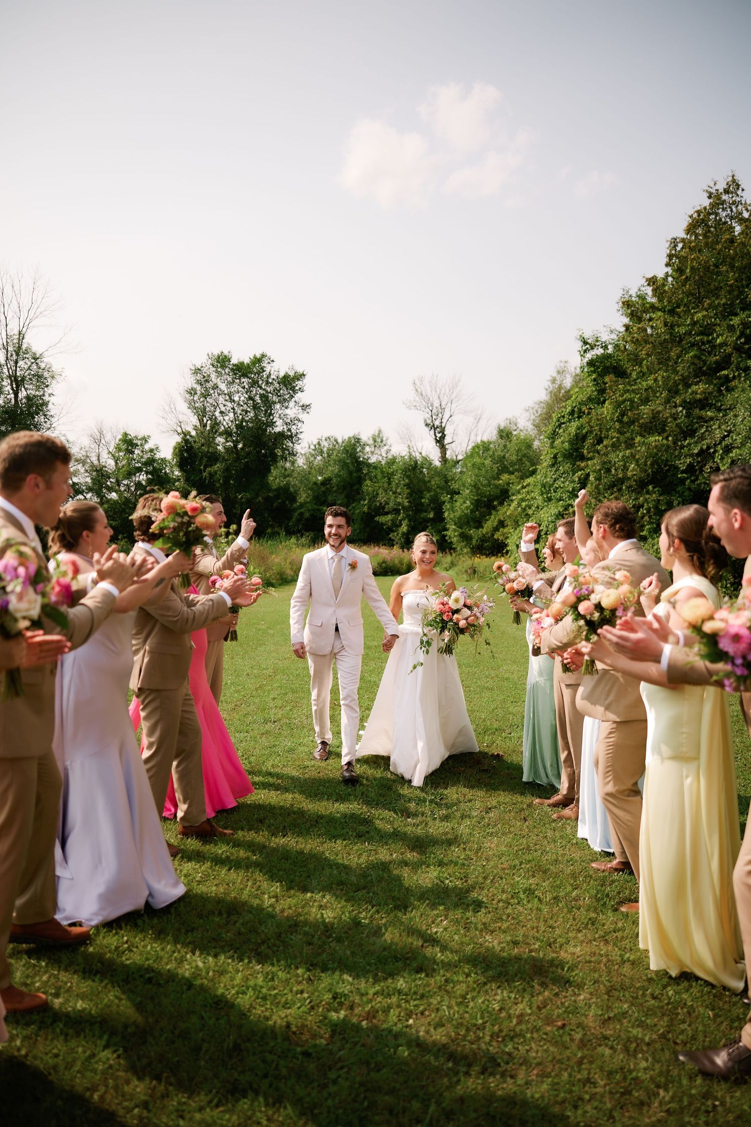 Bride and groom walk through a floral arch formed by colorful bridesmaids and groomsmen on a lush green lawn.
