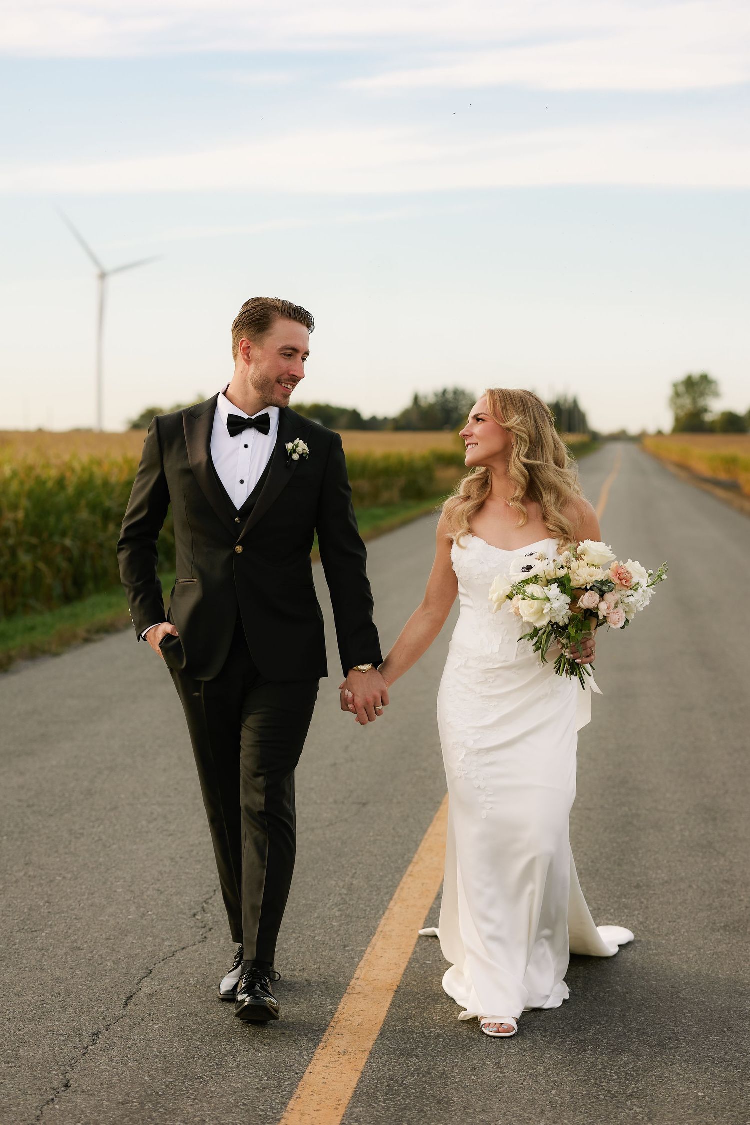 Bride in a white gown and groom in a black tuxedo hold hands while walking down a country road past cornfields and a wind turbine.