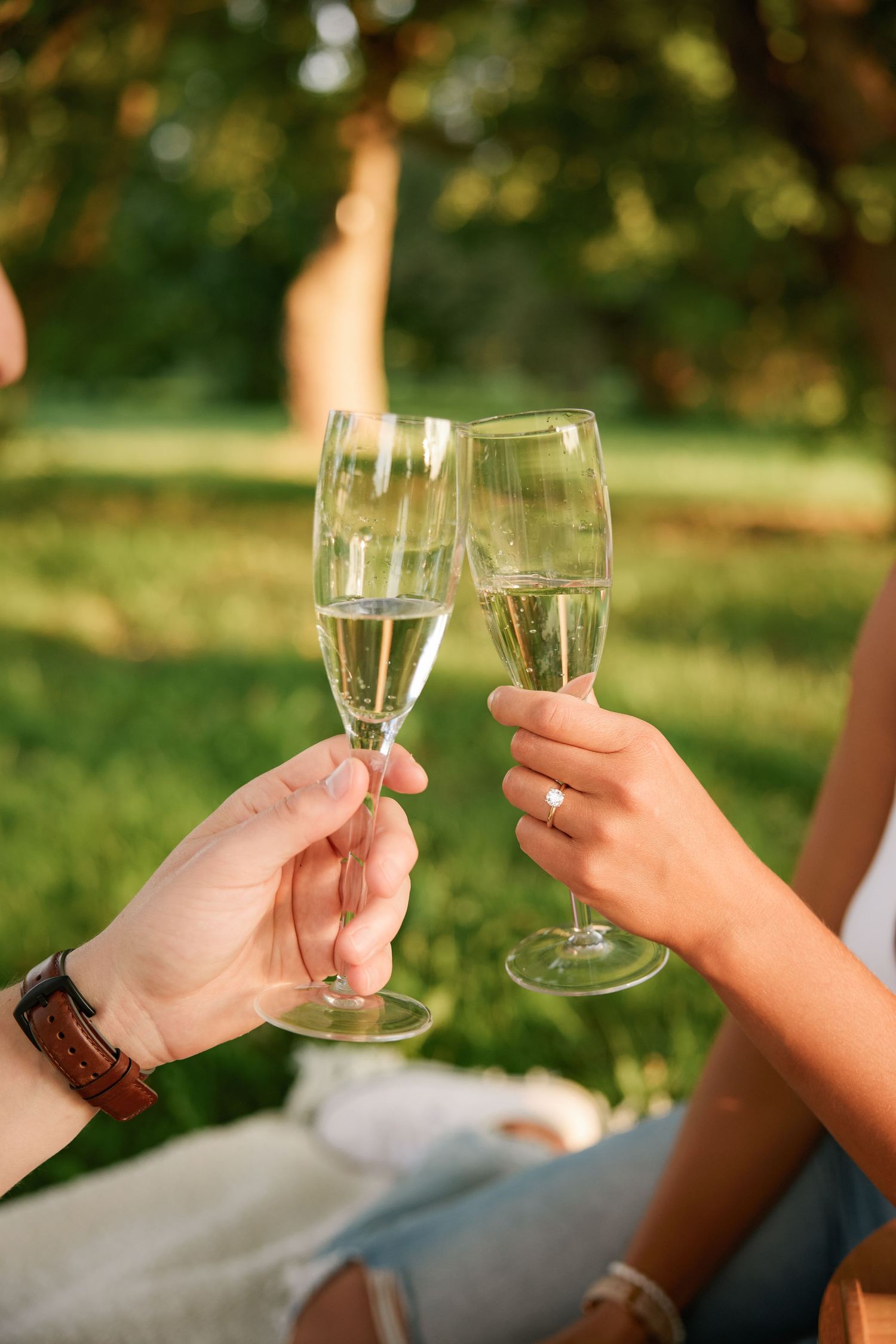 Couple toasting with champagne glasses outdoors, woman wearing an engagement ring celebrating in a green park.