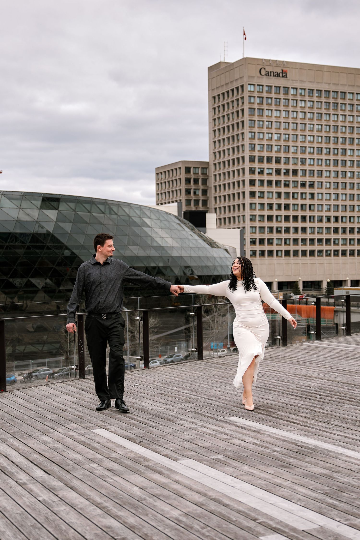 Couple holding hands on wooden deck with Canada government building and glass dome architecture in background.