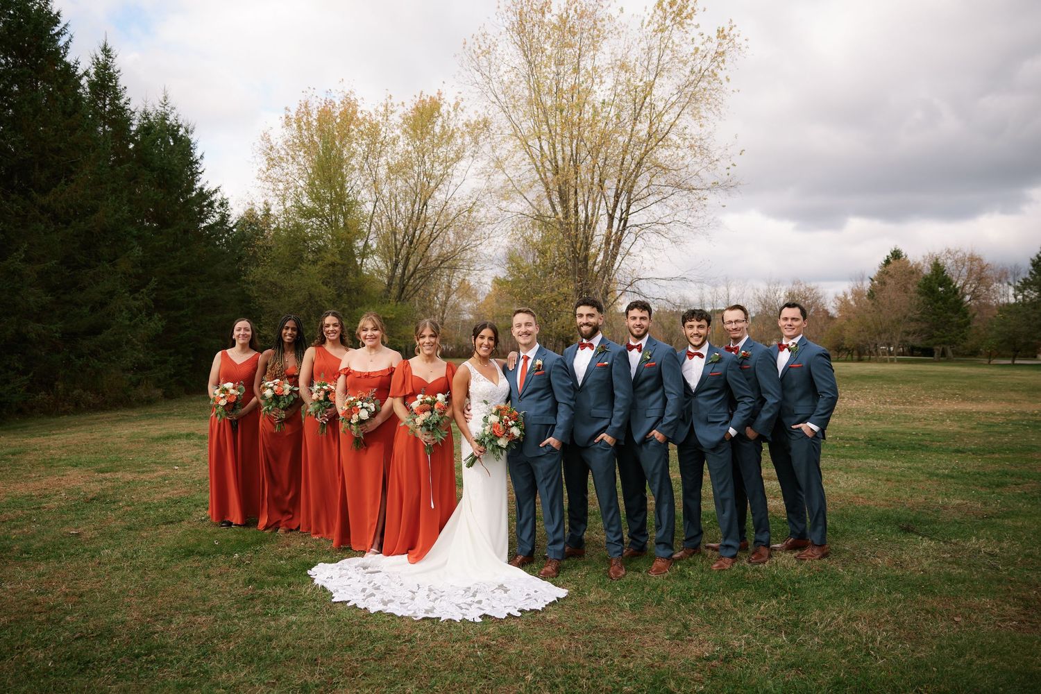 Wedding party poses for photos in a field with bridesmaids in red and groomsmen in navy blue.
