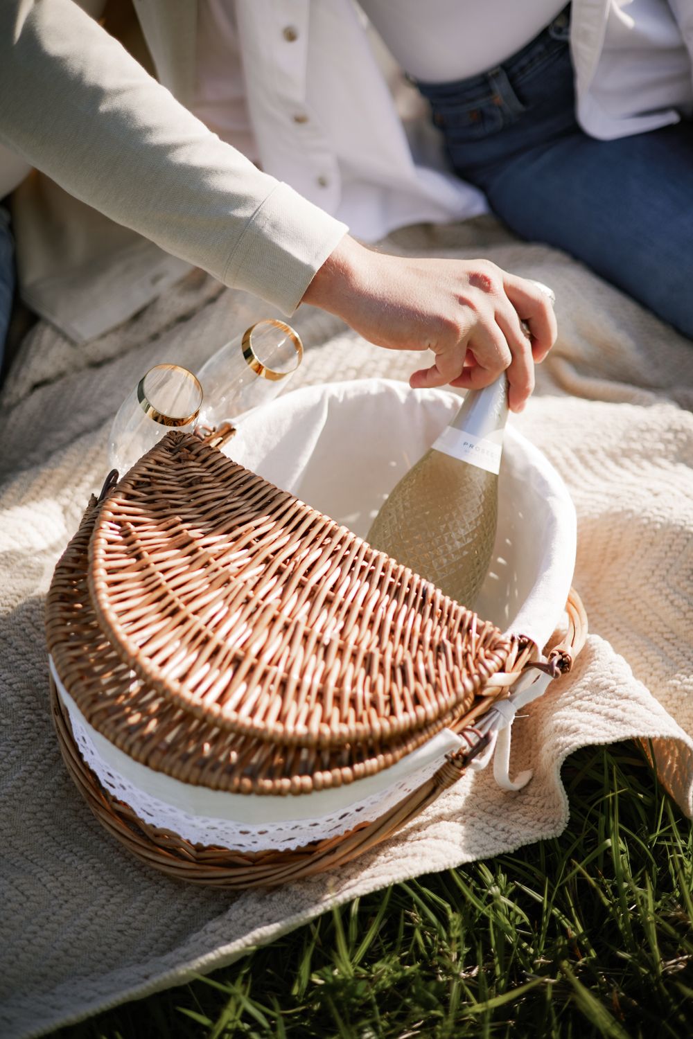 Woman opening a wicker picnic basket on a blanket outdoors, wearing jeans and a gold bracelet.