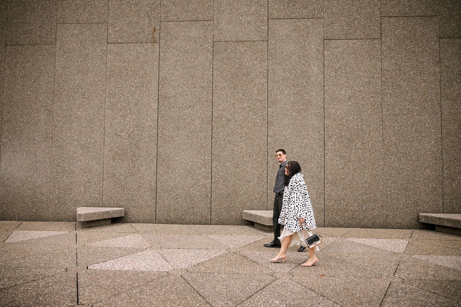 Couple walking past a large granite stone wall, woman in floral dress and man in suit, urban architecture setting.