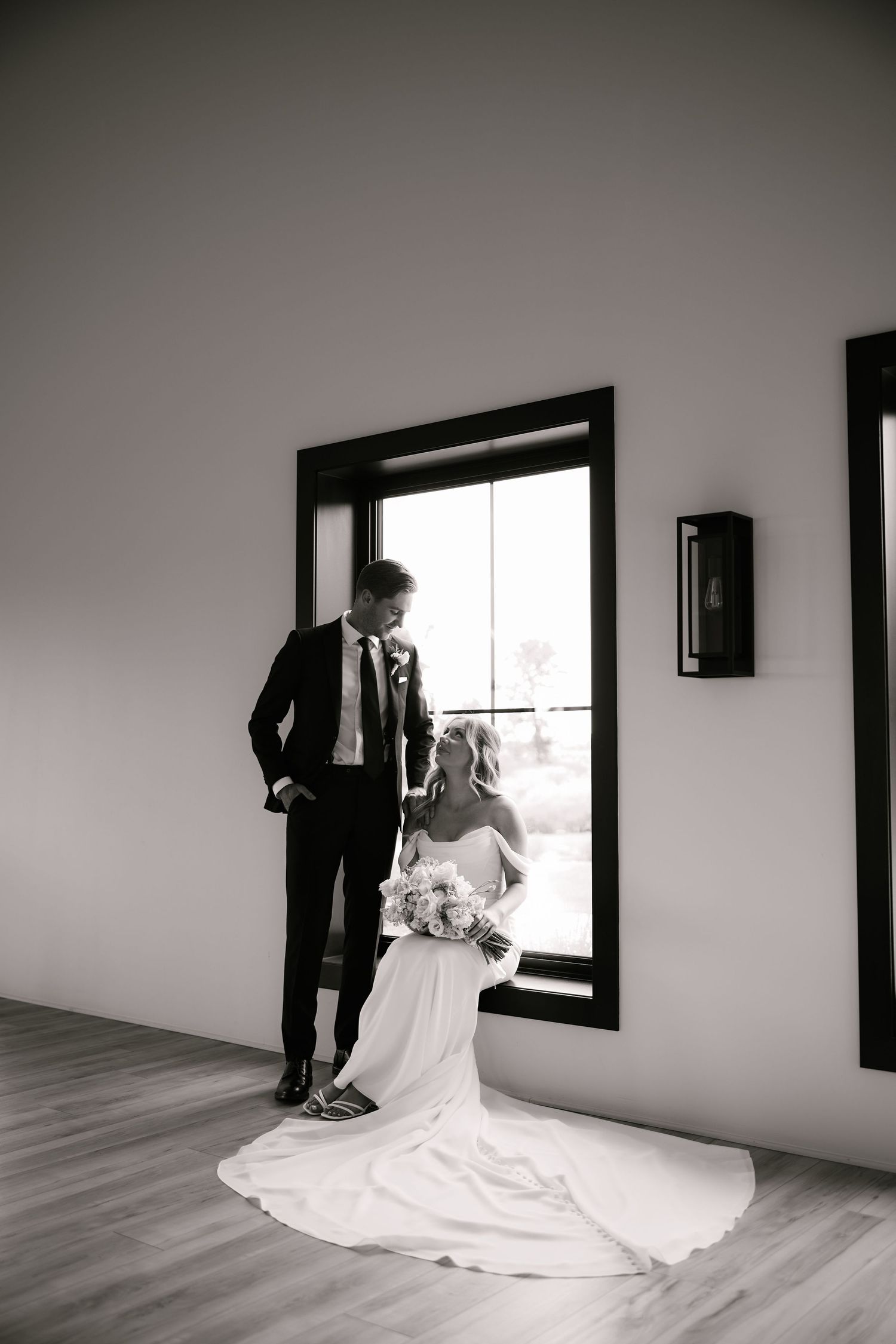 Black and white portrait of bride and groom sharing tender moment by large window in minimalist wedding venue.