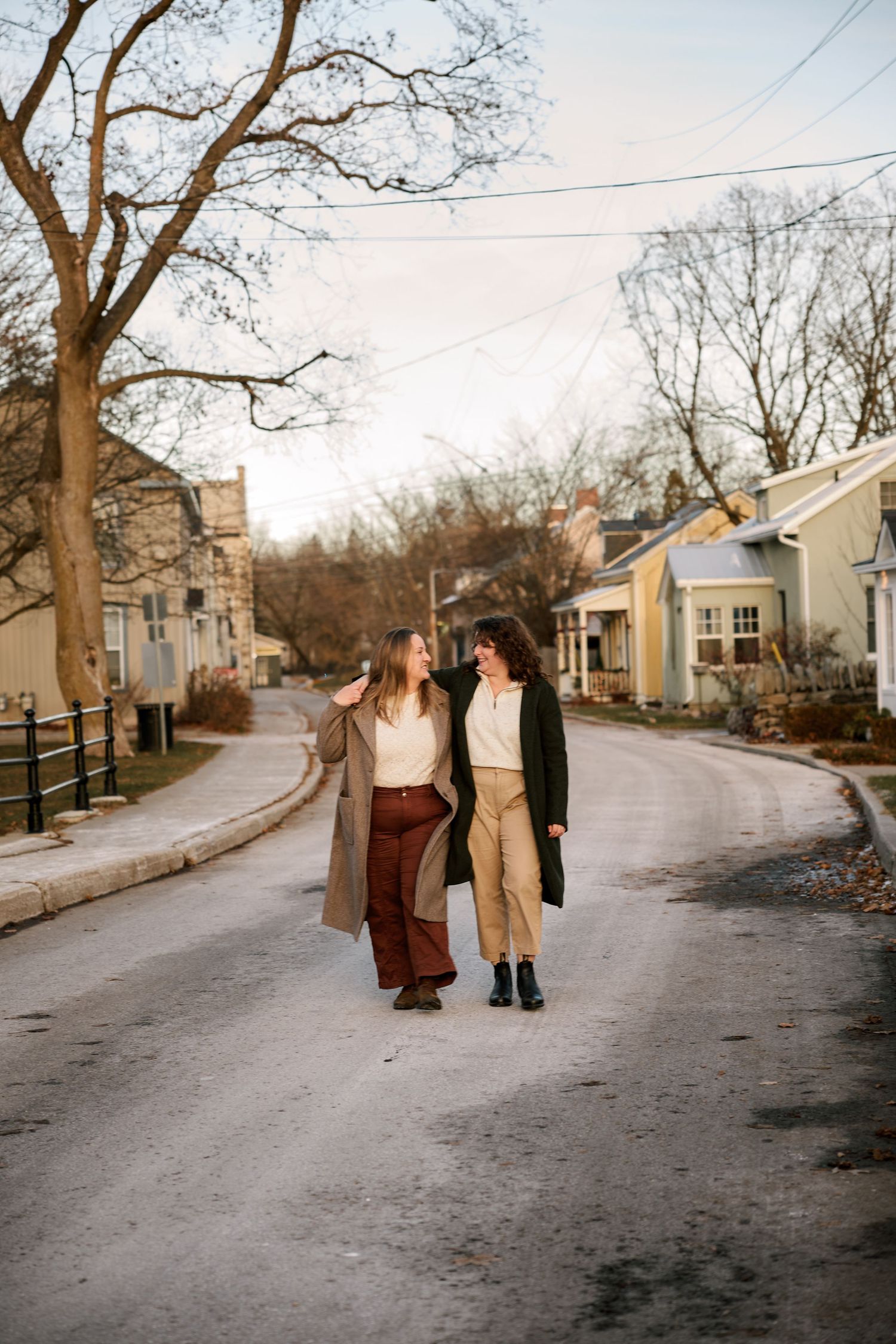 Two women walk together on a quiet residential street in autumn, smiling at each other in long coats.
