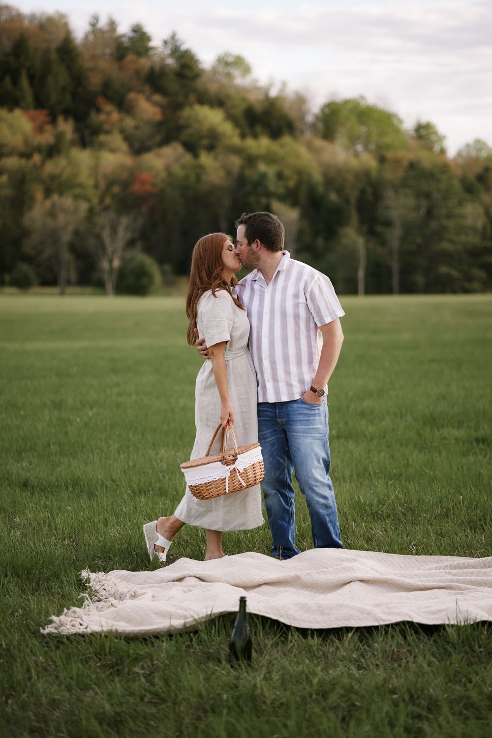Couple sharing a romantic kiss during a picnic in a lush green field, holding a wicker basket with a blanket on the grass.