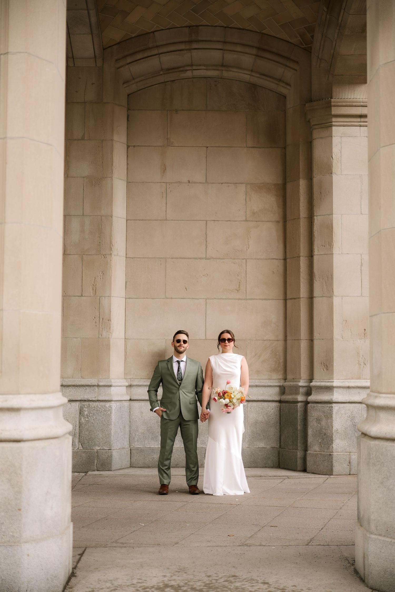 Bride and groom standing together under stone archway columns, bride holding colorful bouquet, groom in sage green suit.